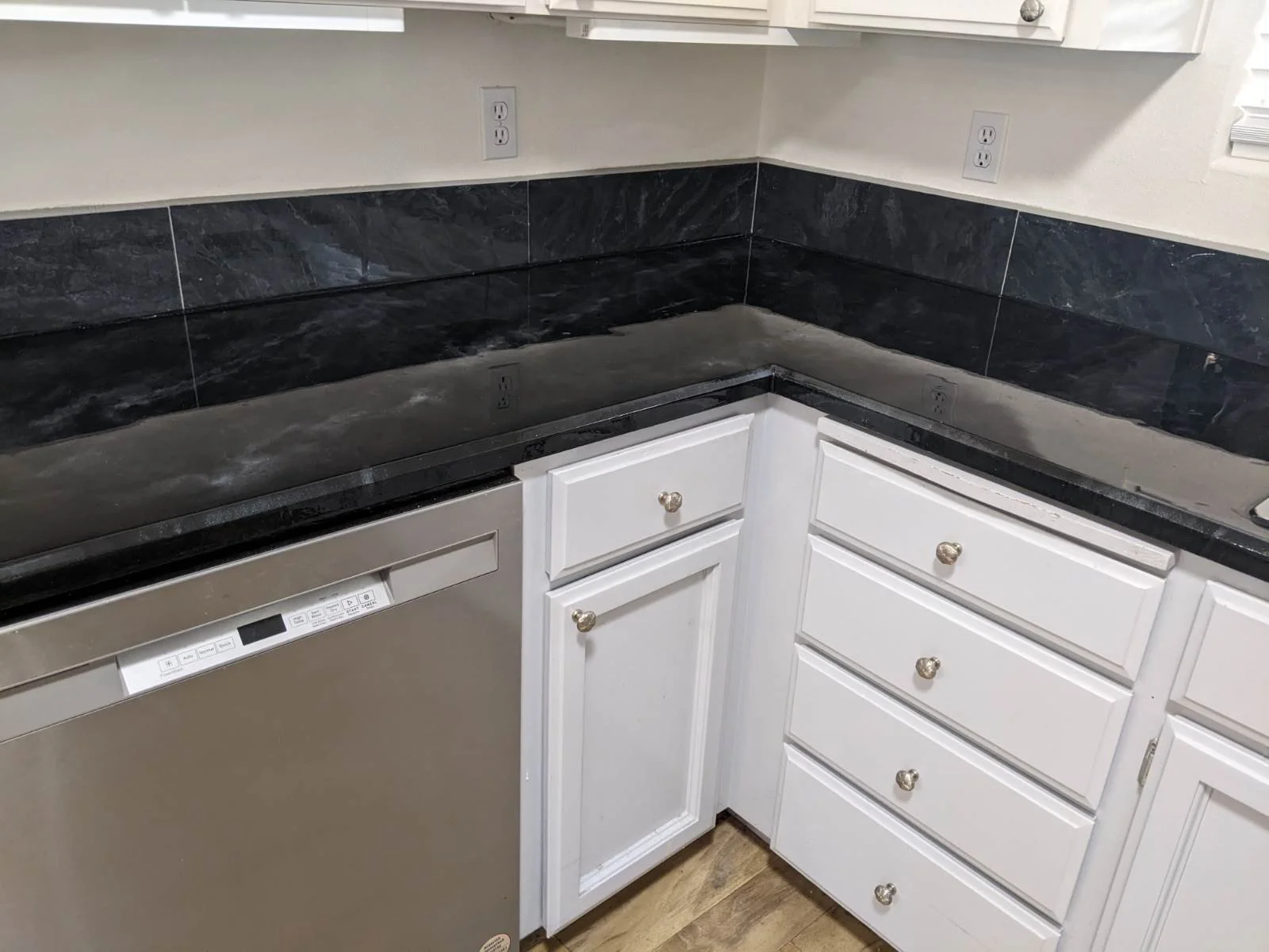Kitchen corner with black marble countertop, white cabinets, dishwasher, and electrical outlets.