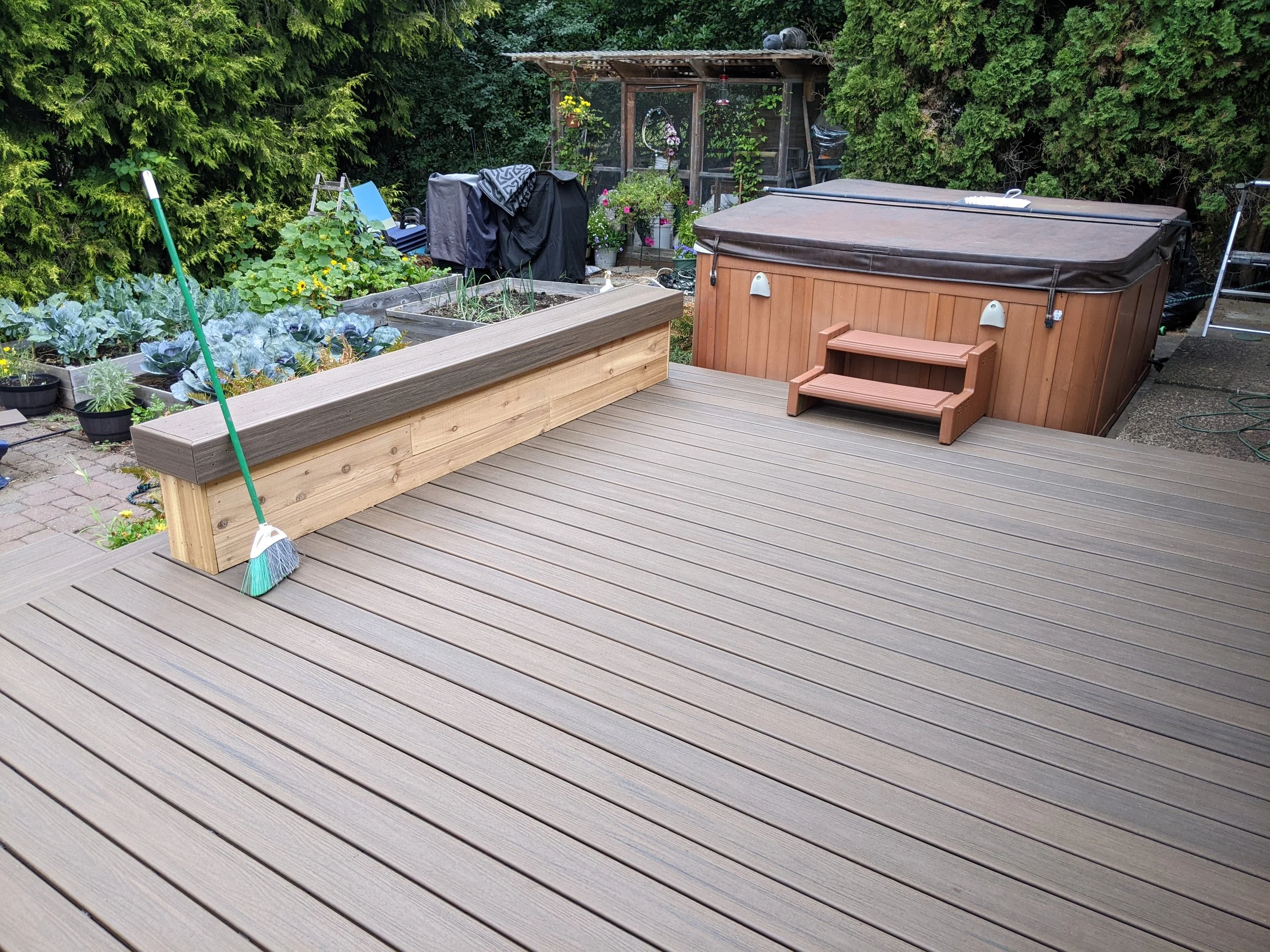 Backyard deck with a hot tub, a potted plant, and a garden bed with vegetables and flowers, surrounded by green trees.