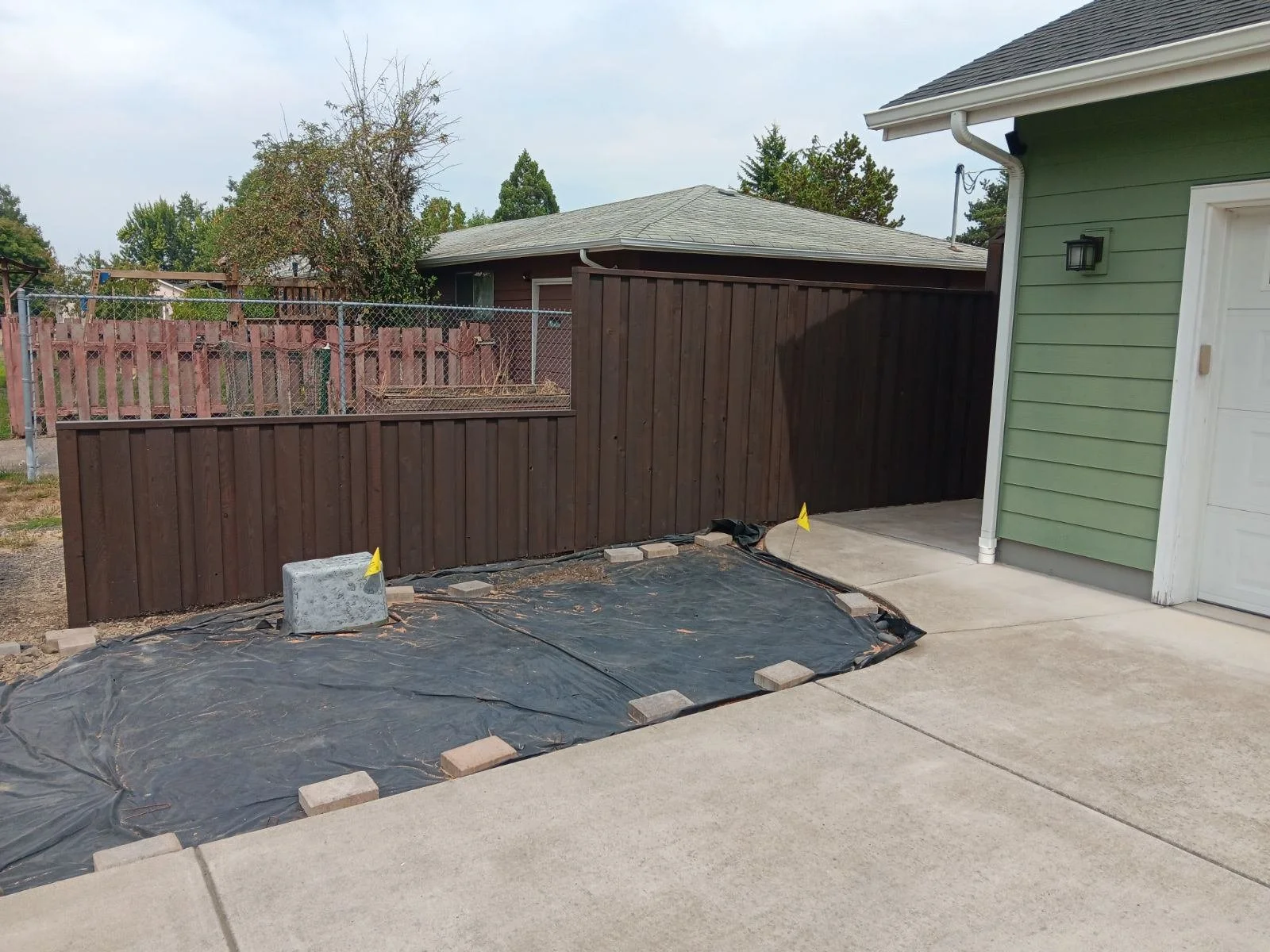 A backyard with a new brown wooden fence under construction, a black tarp on the ground with bricks around the edges, and a light green house with white trim and a white garage door.