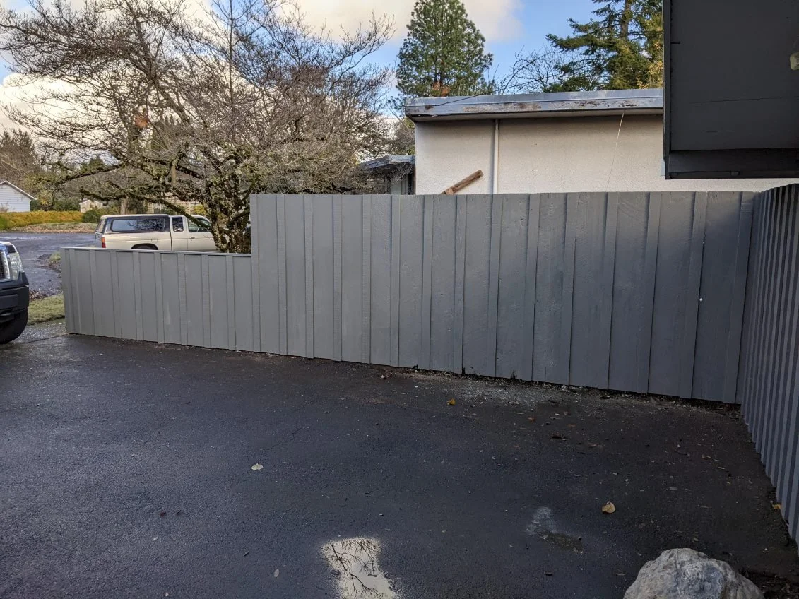Gray wooden fence along a driveway with trees and a house in the background.