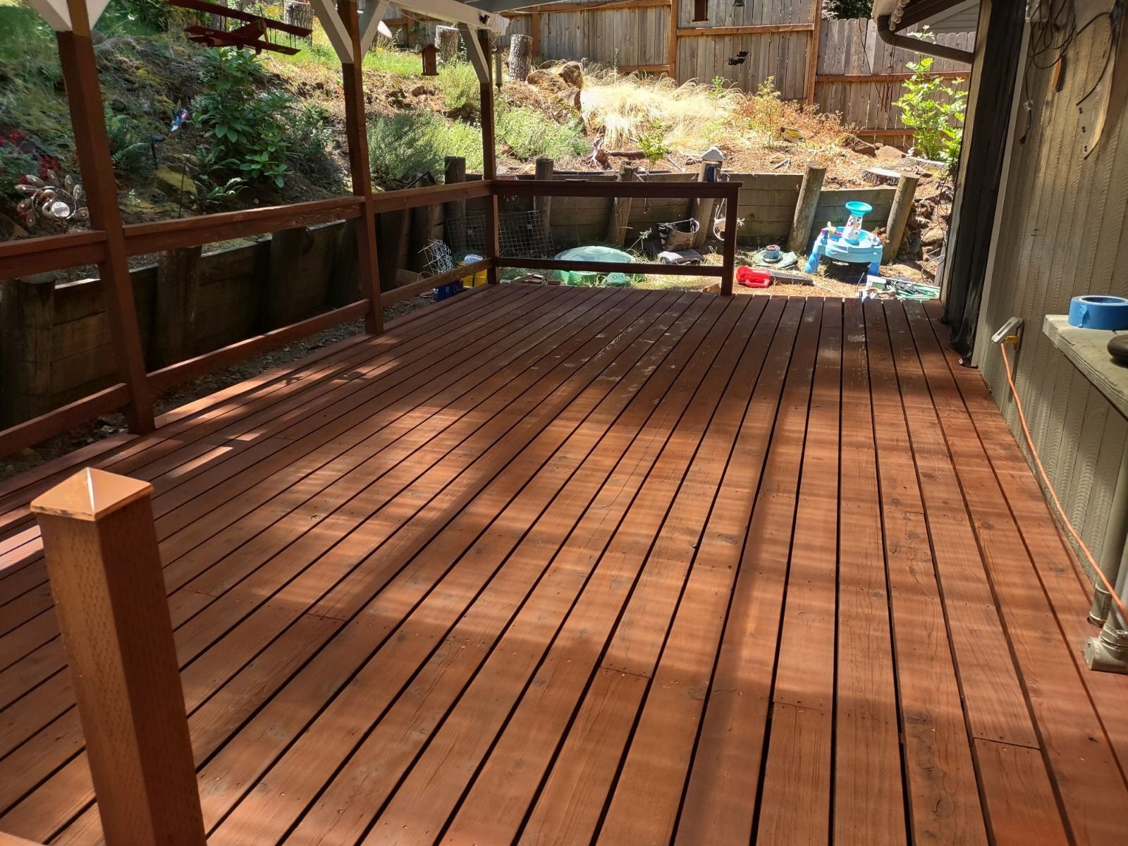 Freshly built wooden deck attached to a house, with a view into an outdoor yard filled with children's toys and garden tools, surrounded by a wooden fence and lush green plants.