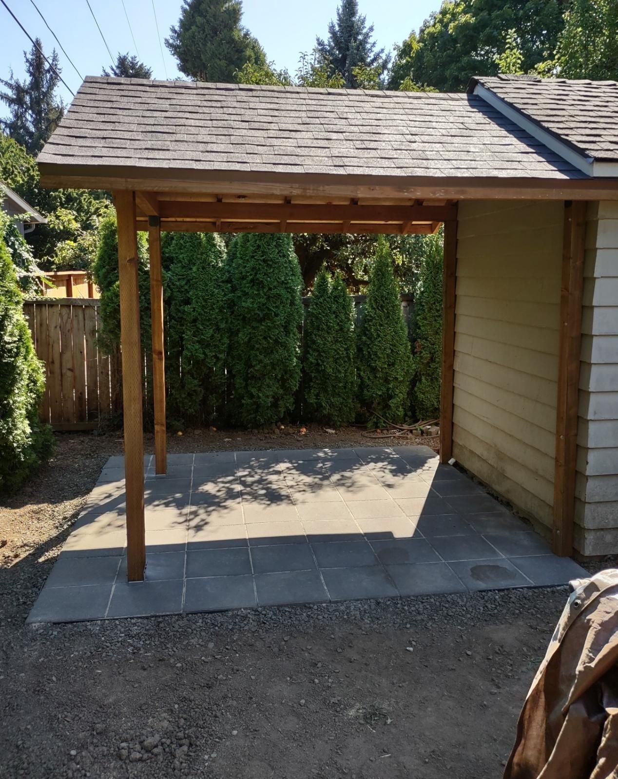 A newly built wooden carport with a shingled roof, standing over a tiled concrete pad next to a house, with a background of green trees and shrubs.