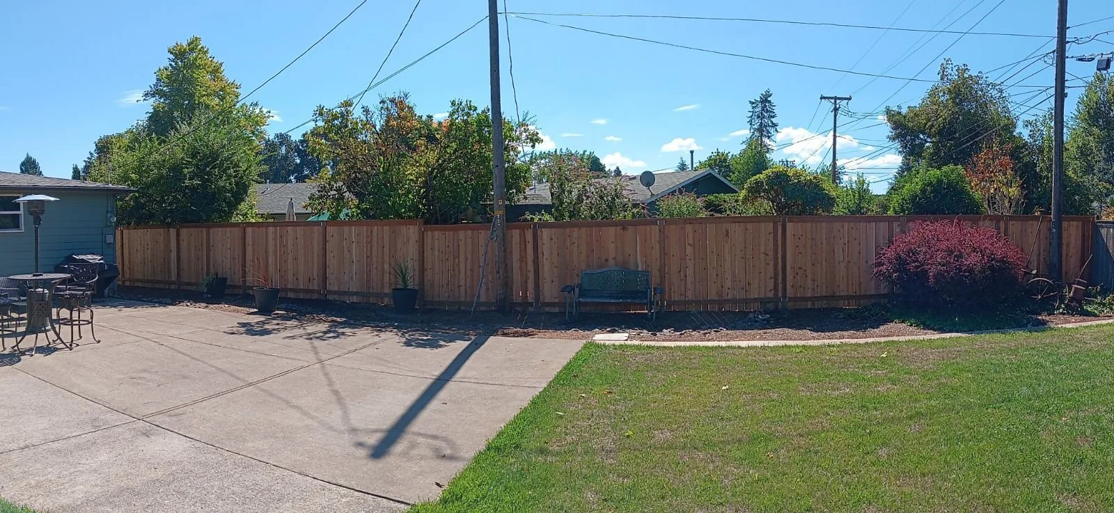 Backyard with a wooden fence, patio furniture, a bench, potted plants, a grassy area, trees, and power lines under a partly cloudy sky.