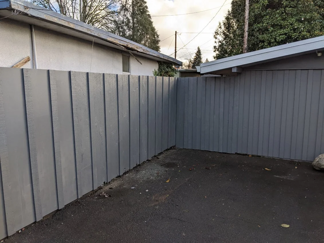 Gray metal fence in a backyard with a dark asphalt surface and trees in the background.