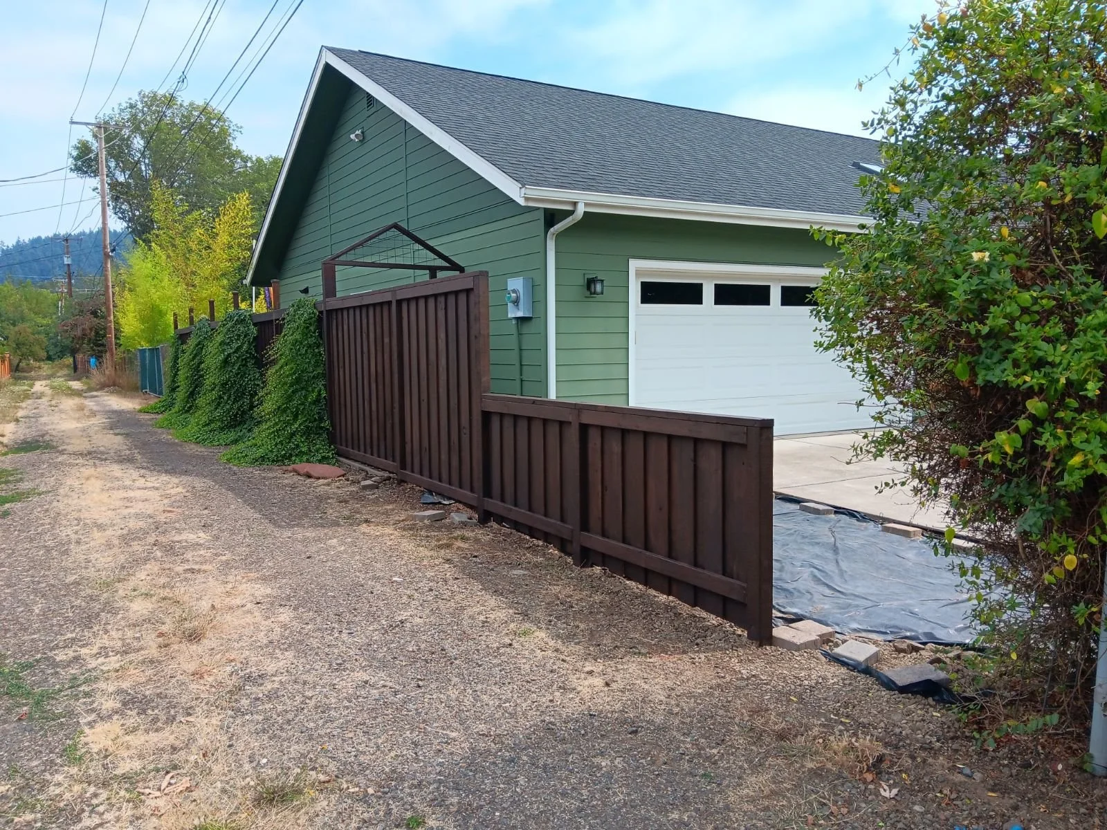 A green house with a white garage door, surrounded by a wooden fence and greenery, on a gravel road under a blue sky.