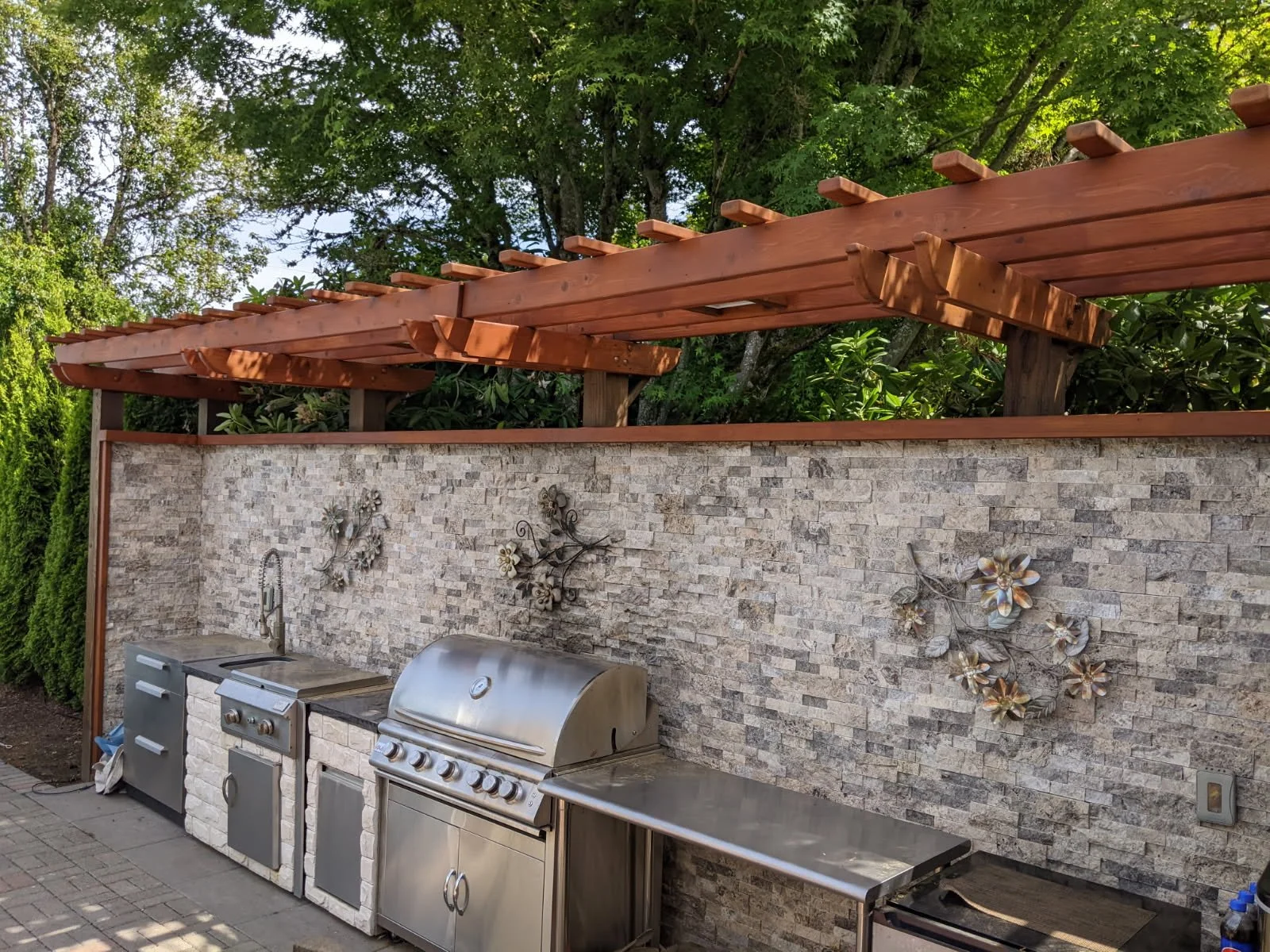Outdoor kitchen area with stone wall, metal grill, small countertop, and wall decorations, surrounded by greenery and a wooden pergola.