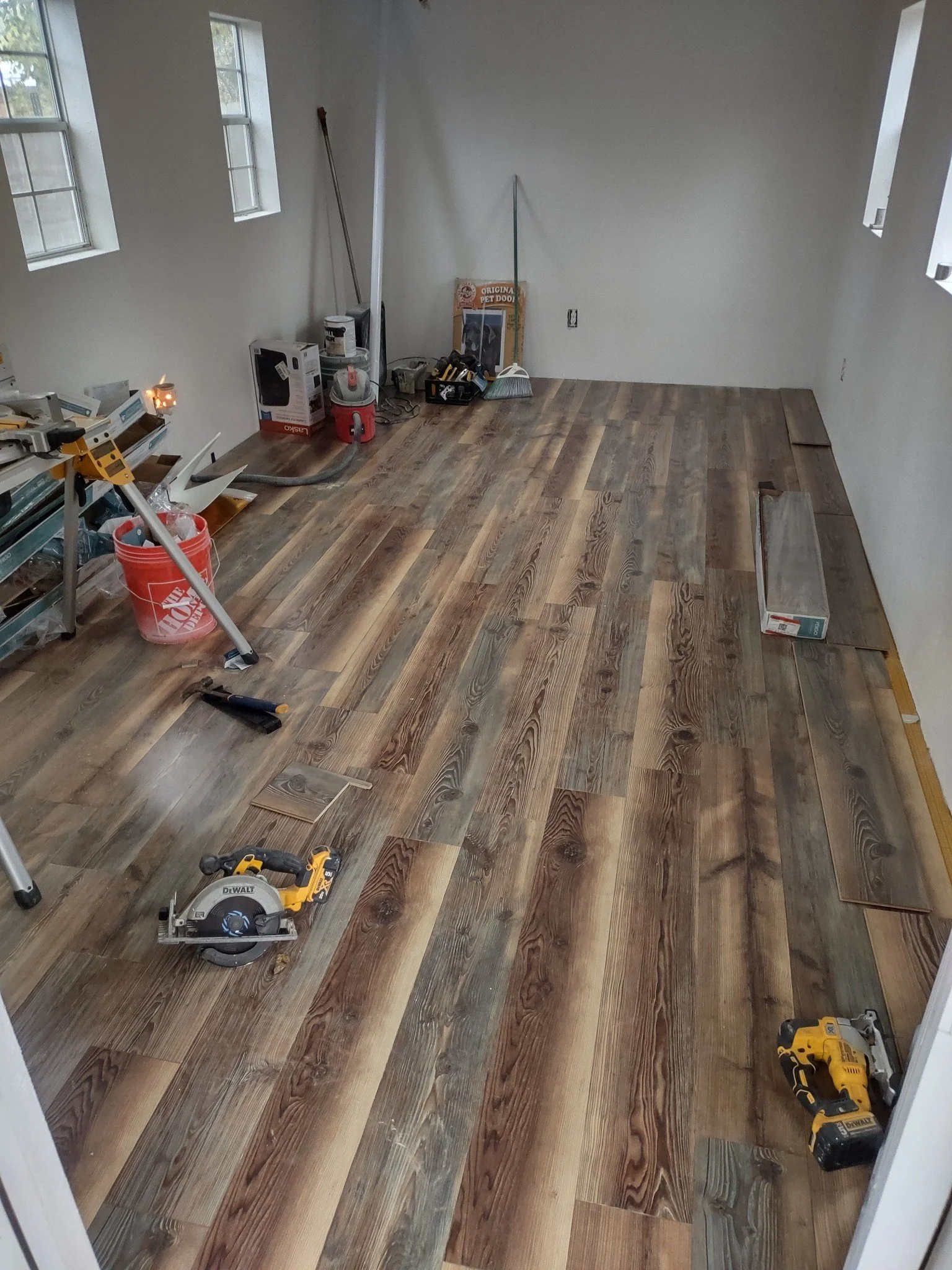Room with wood flooring in progress, tools scattered, including a saw, drill, and measuring tape, with windows on the left and some boxes and materials in the back corner.