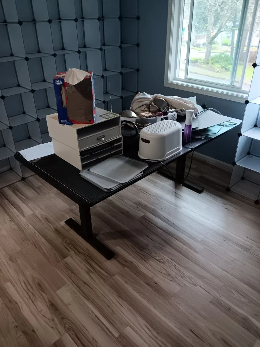 Messy black table against a gray wall with square 3D patterns, cluttered with various items including a box of tissues, a small white drawer organizer, papers, a white and purple ultrasonic diffuser, a hairdryer, and a cloth near a window with a view