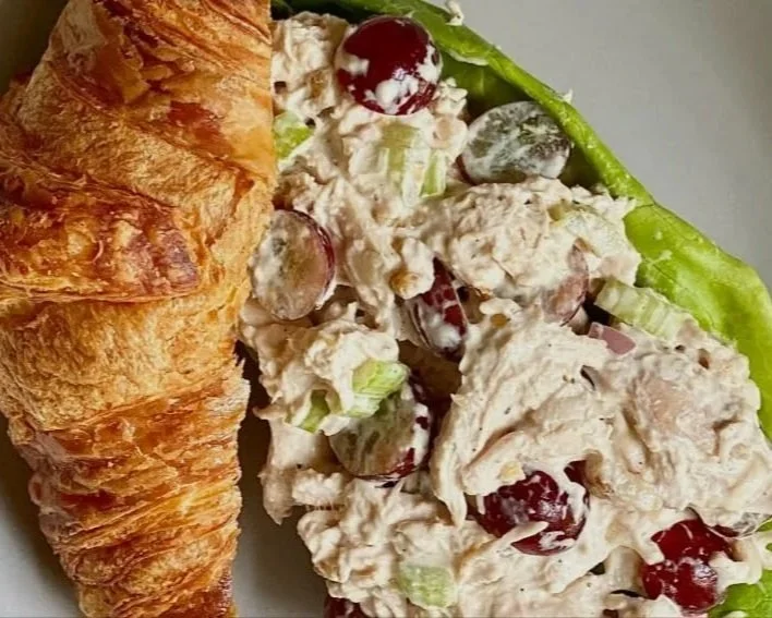 A plate with a croissant and a serving of chicken salad with grapes and celery, garnished with lettuce and a cherry on top.