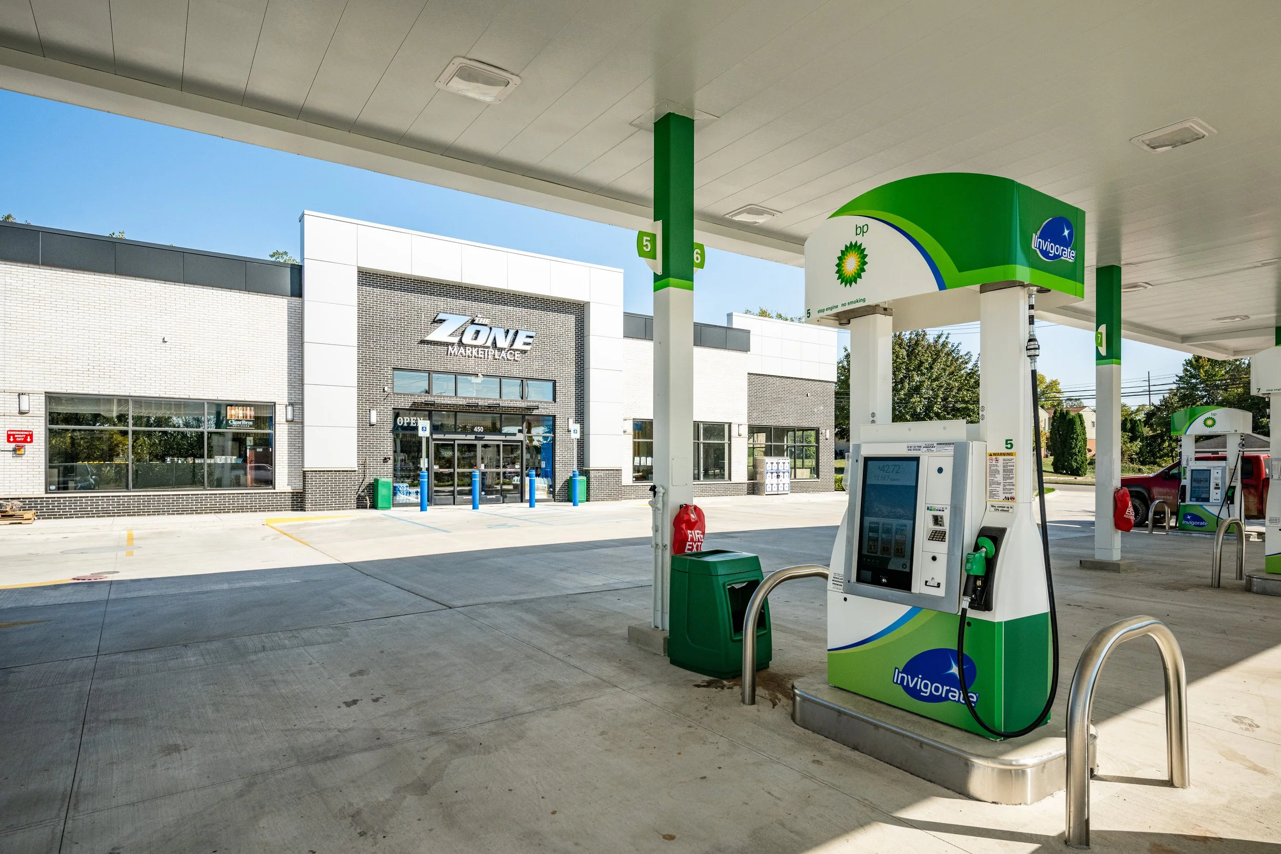 A BP gas station with fuel pumps and a convenience store named "Zone Marketplace" in the background. The gas station is labeled with numbers 5, 6, and 7 for the fuel pumps. Clear blue sky and some trees are visible in the background.