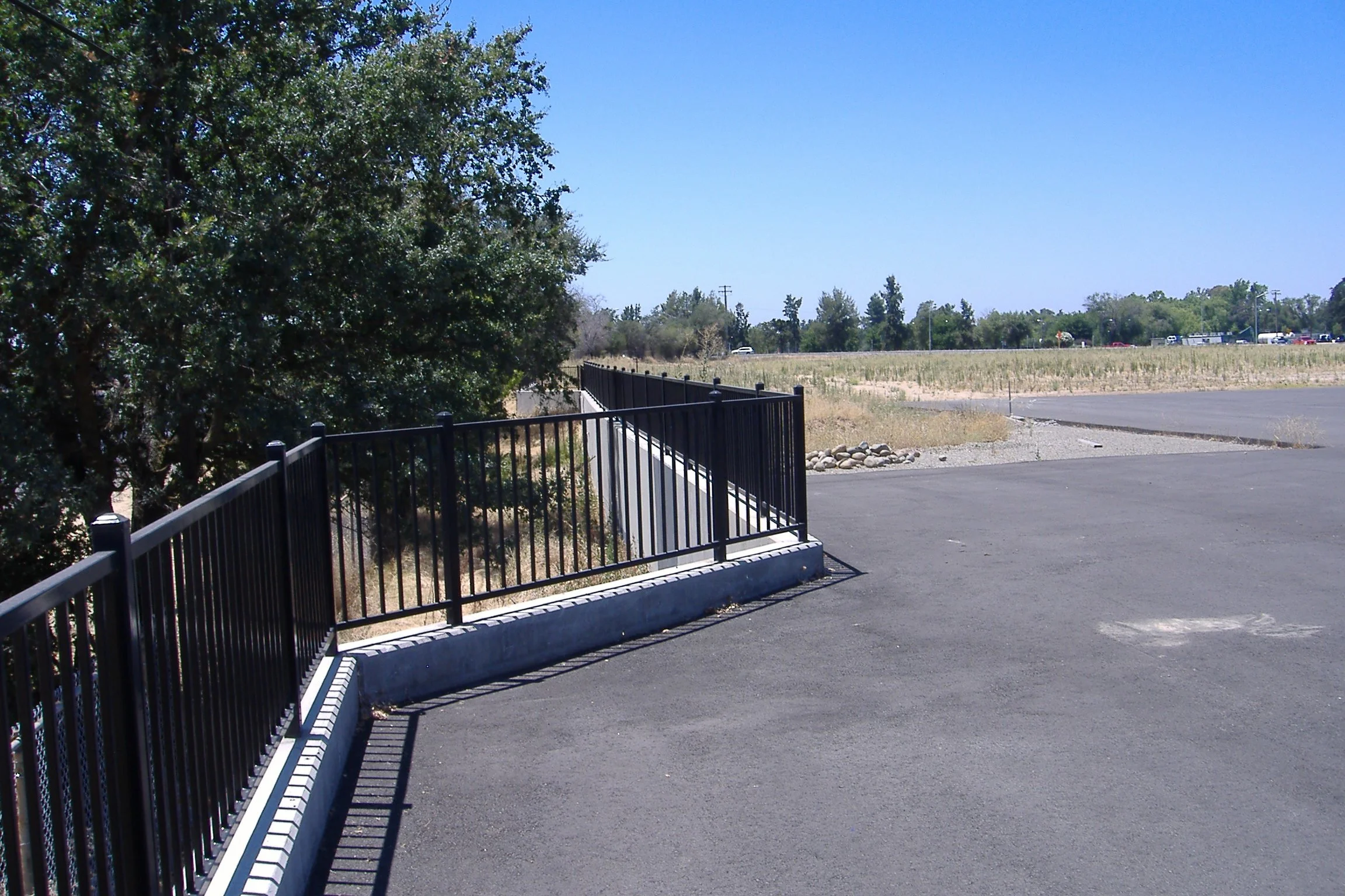A view of a black metal fence along a concrete sidewalk with a large green tree on the left, a dirt grass field in the background, and a parking lot with cars under a clear blue sky.