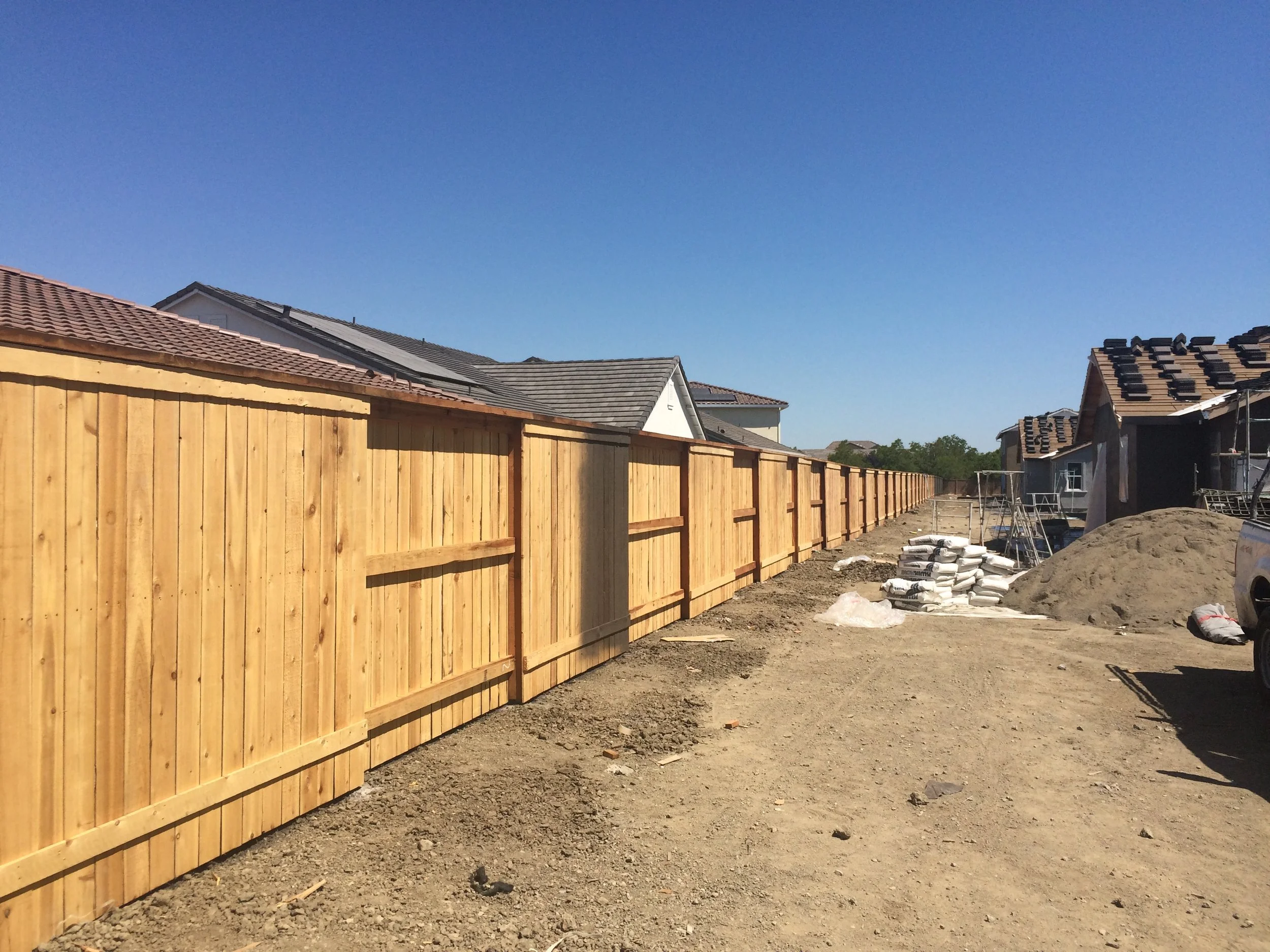 Construction site showing a long wooden fence, partially built houses with rooftops, scaffolding, and construction materials on the ground under a clear blue sky.