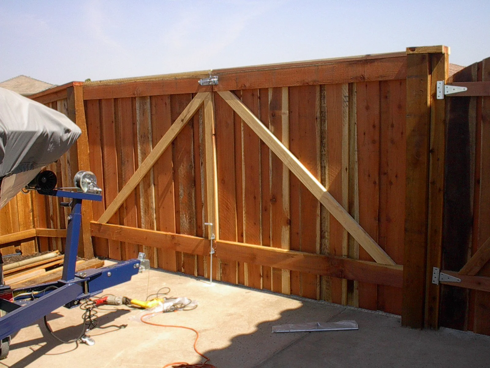 A wooden fence with a gate under construction, supported by diagonal braces, in a backyard. There is a piece of equipment and some tools on the ground.