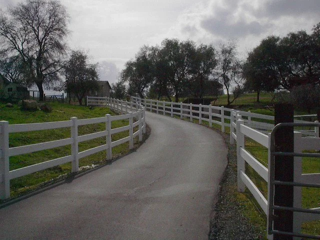 A paved country road with white fence on both sides, curved and leading into the distance among trees and grass under a cloudy sky.