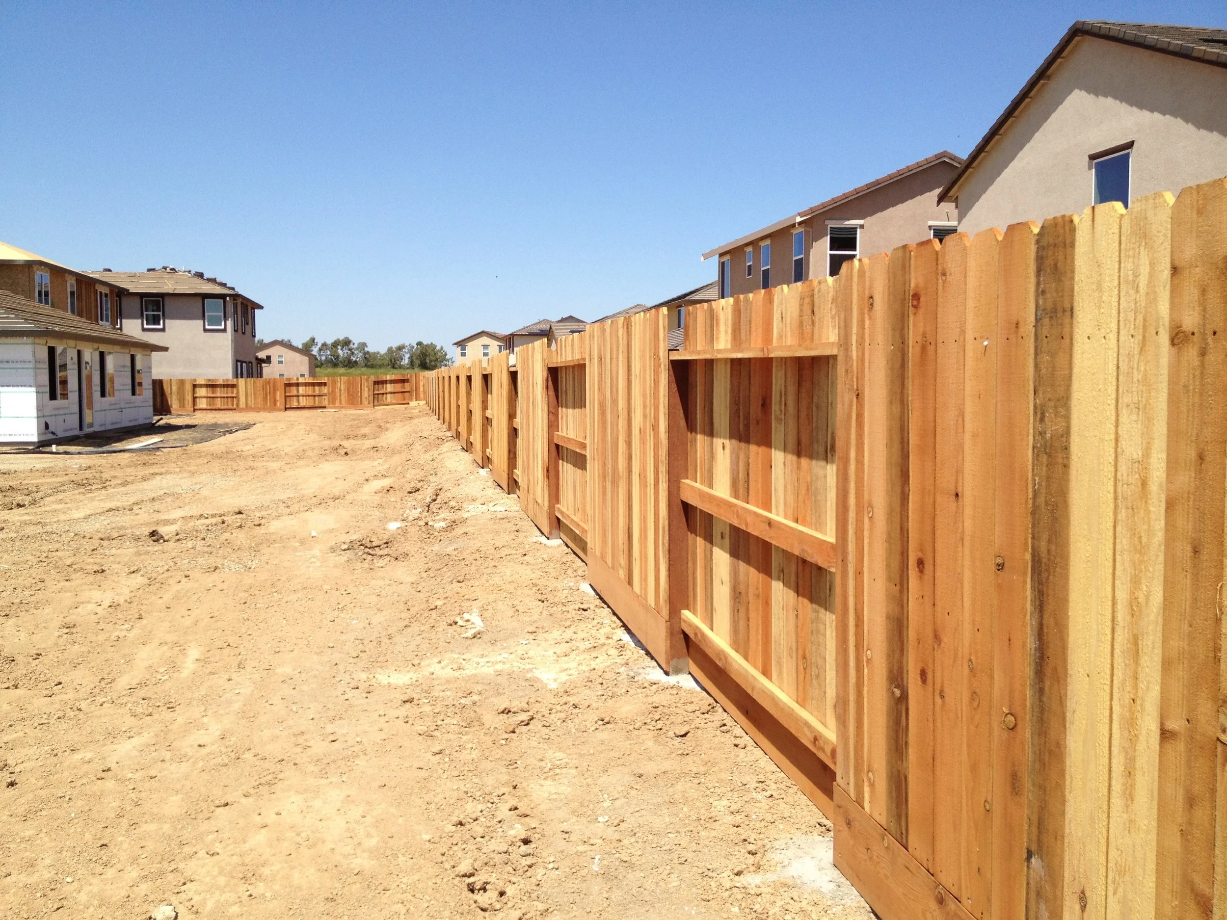 View of a new residential construction site with a wooden fence, partially built houses, and a clear blue sky.