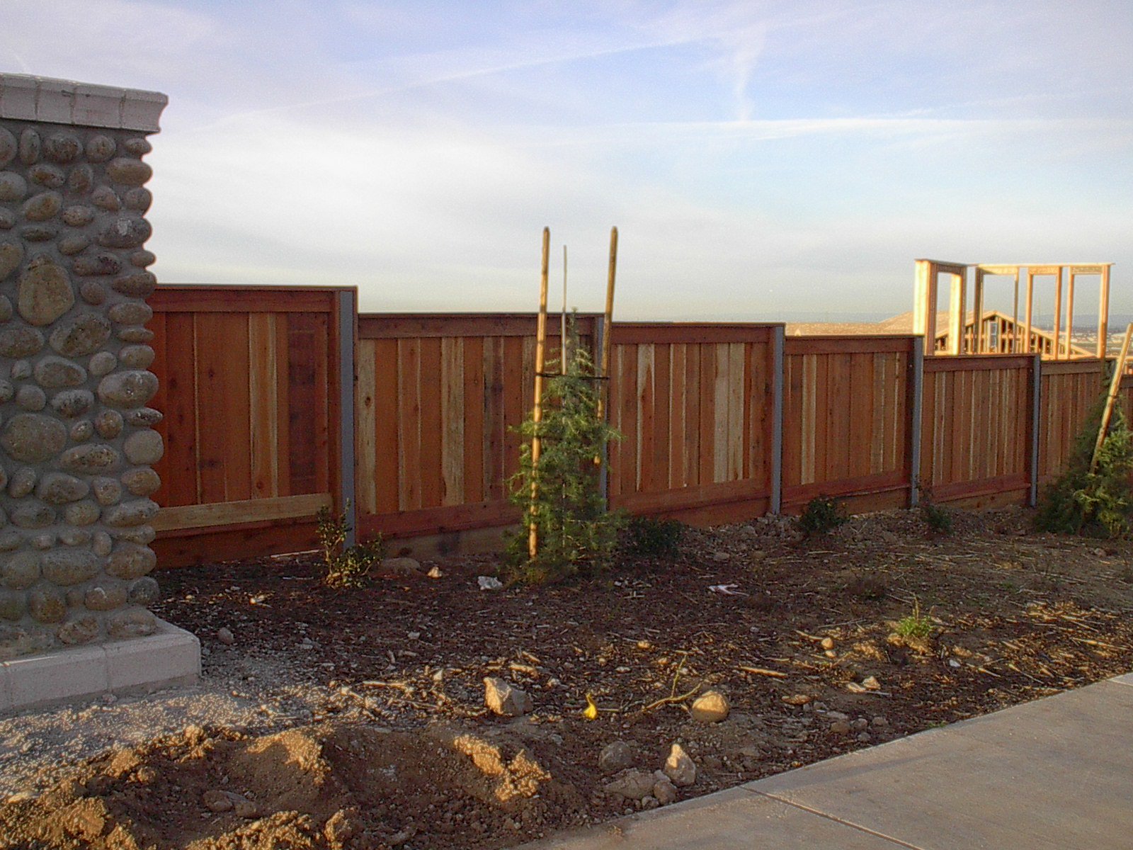A wooden fence under construction in a backyard, with a stone pillar on the left, some young plants supported by stakes, and a blue sky with clouds in the background.