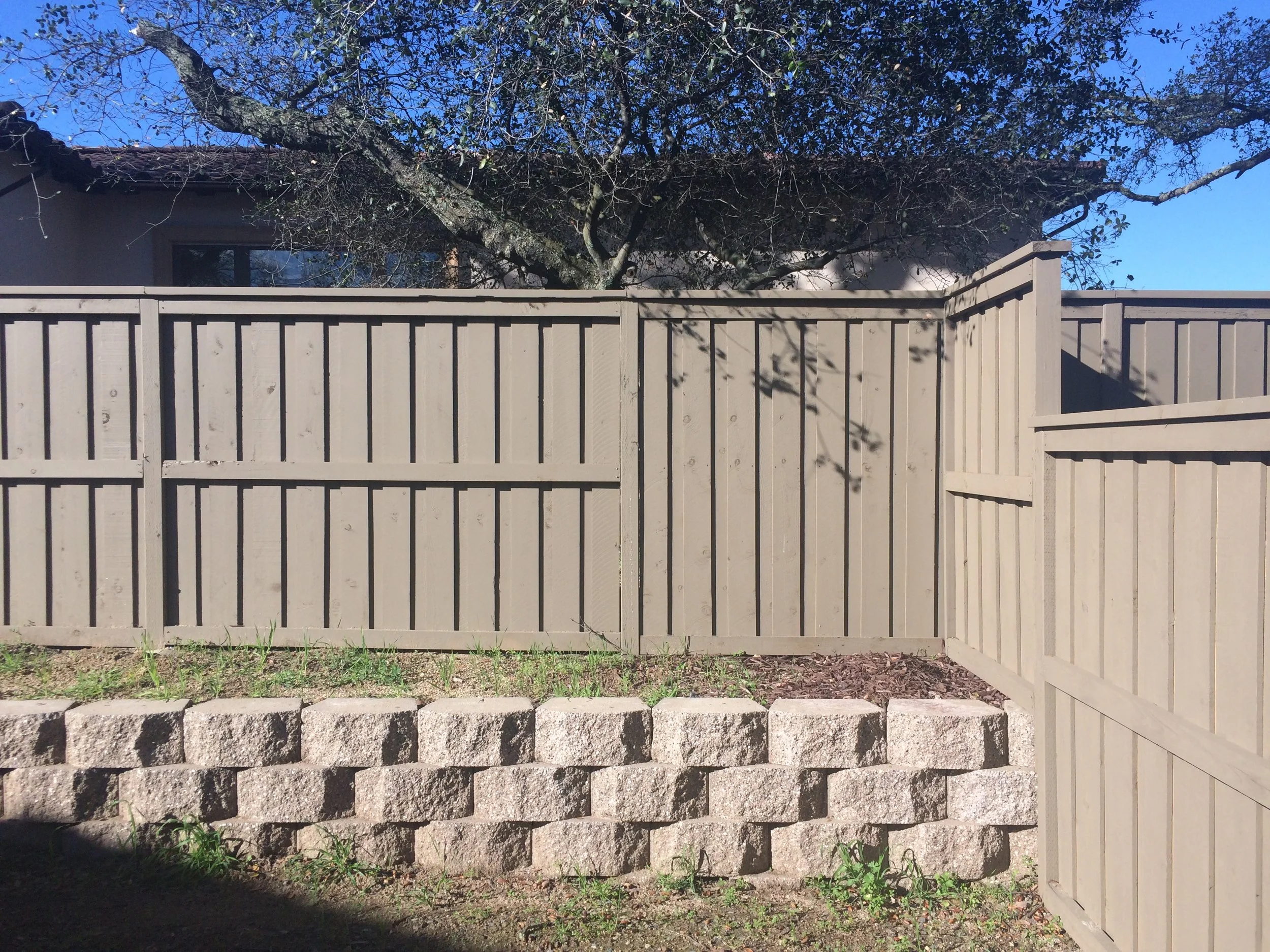 A view of a backyard with a tall tree, a beige fence, a stone retaining wall, and a small patch of grass and dirt.