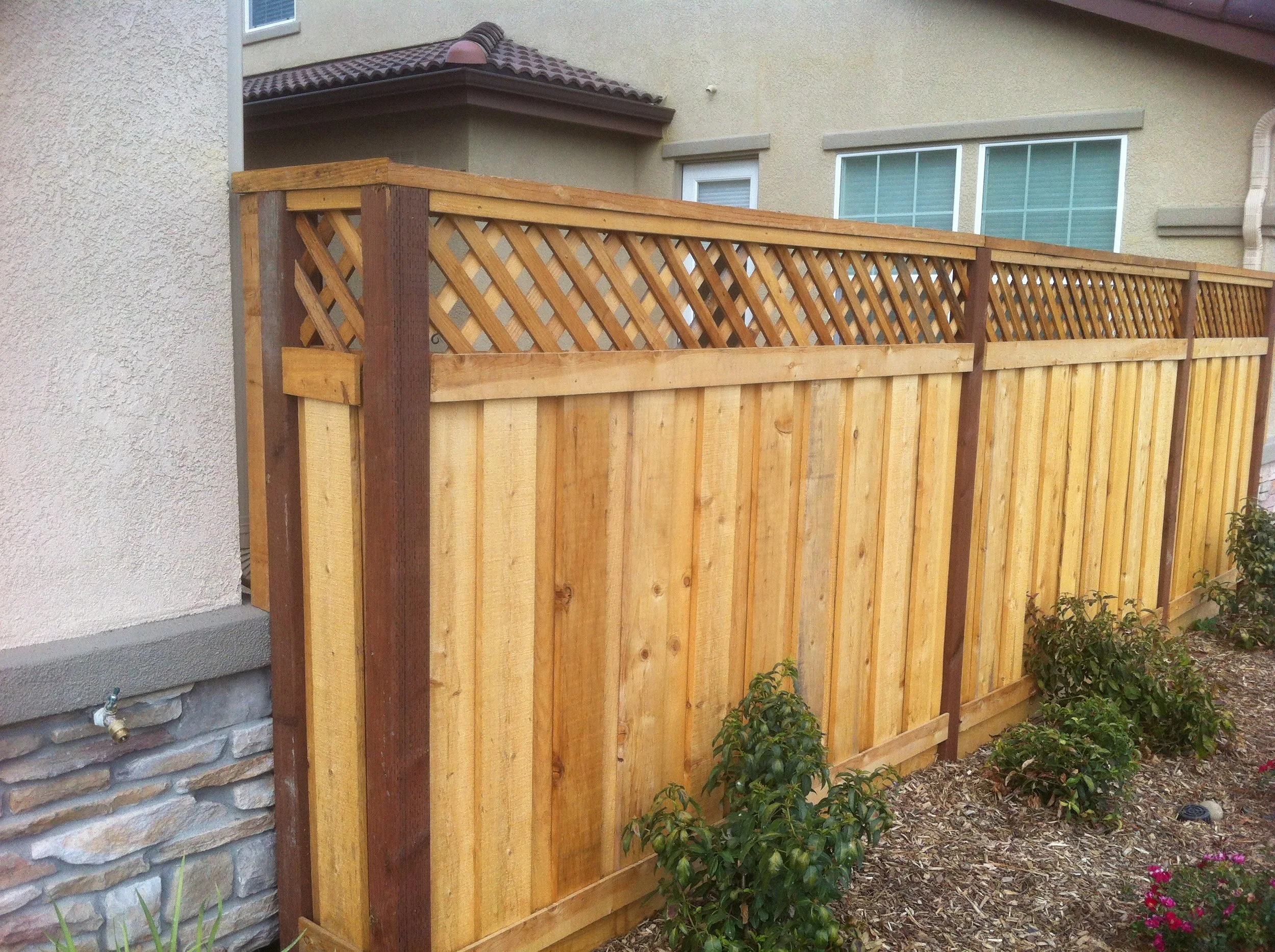 A newly installed wooden privacy fence next to a house with a stucco exterior and a window, with plants and flowers in the garden bed at the base.
