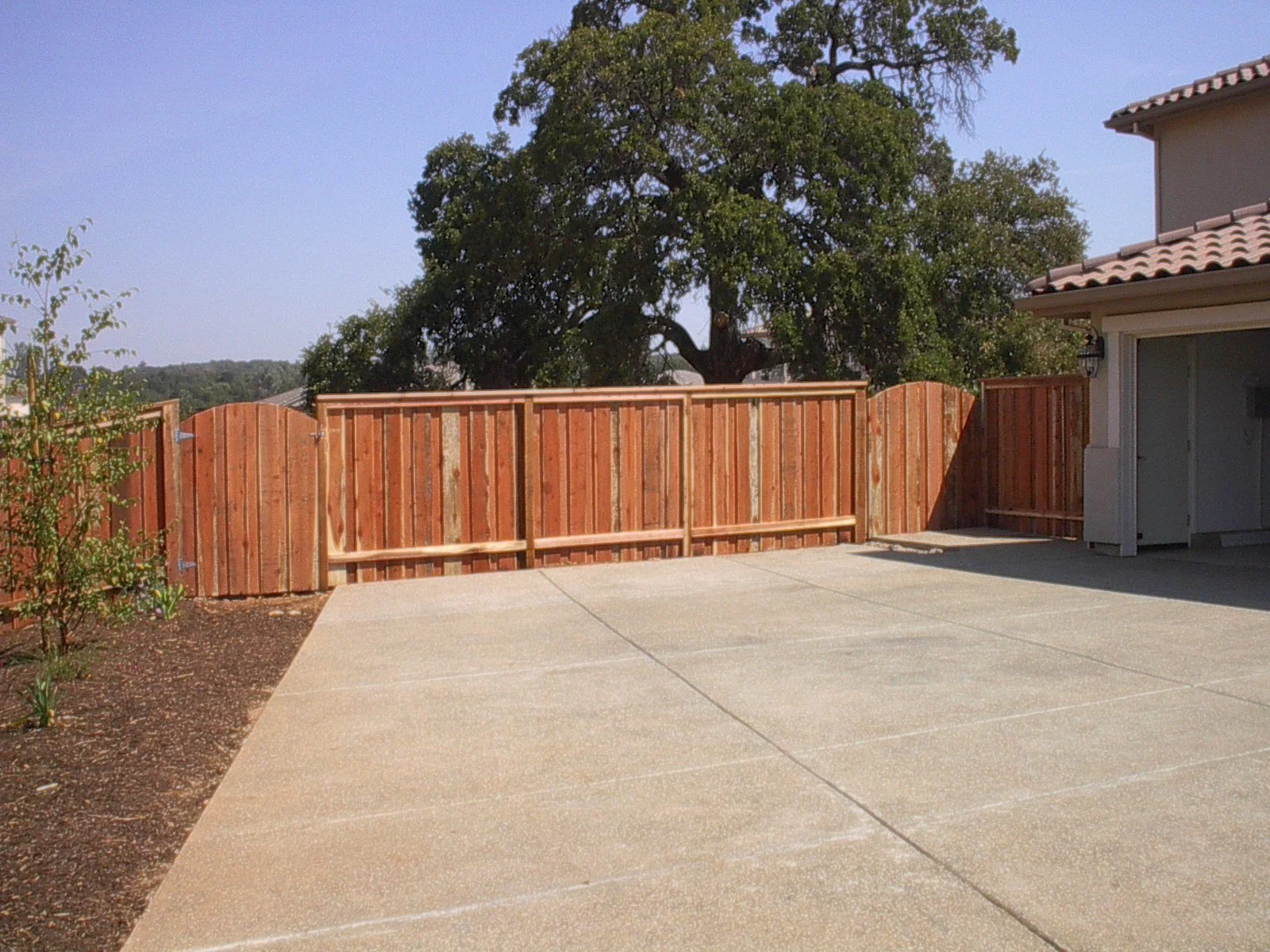 Residential backyard patio with a concrete surface, a wooden fence, tree in the background, and part of a house with a garage on the right.
