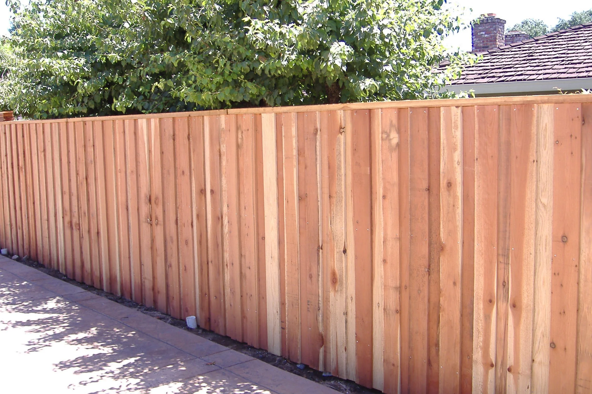 A newly installed wooden fence along a sidewalk with greenery and part of a house roof visible in the background.