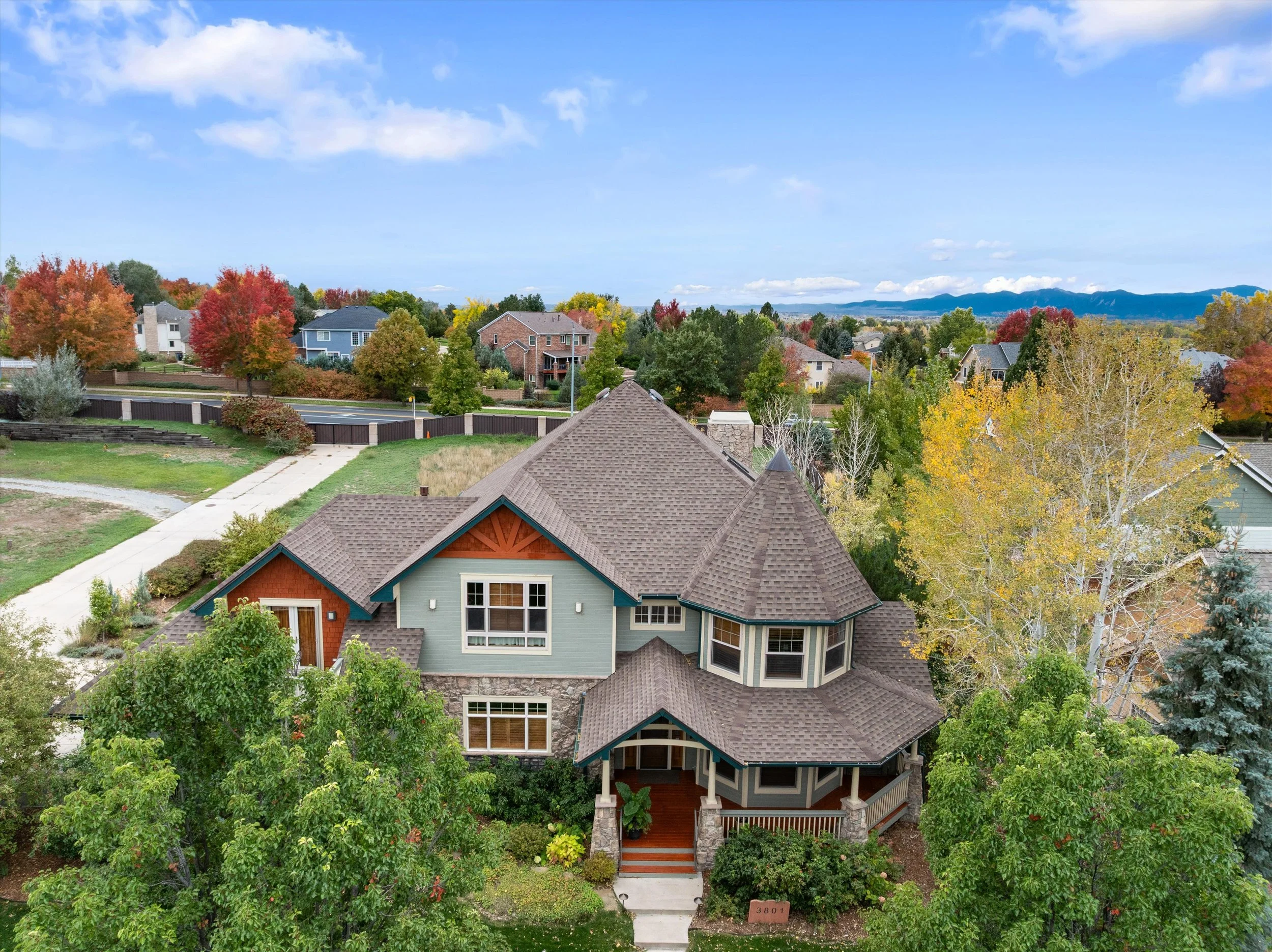 A large, multi-story house surrounded by trees with autumn foliage, in a suburban neighborhood with other houses and distant mountains in the background.