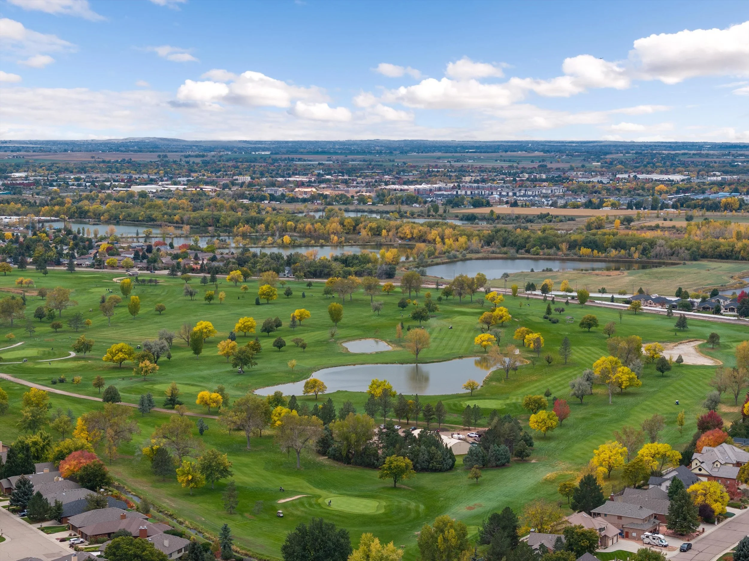Aerial view of a golf course with green fairways, ponds, trees with autumn foliage, nearby houses, and a cityscape in the distance under a partly cloudy sky.