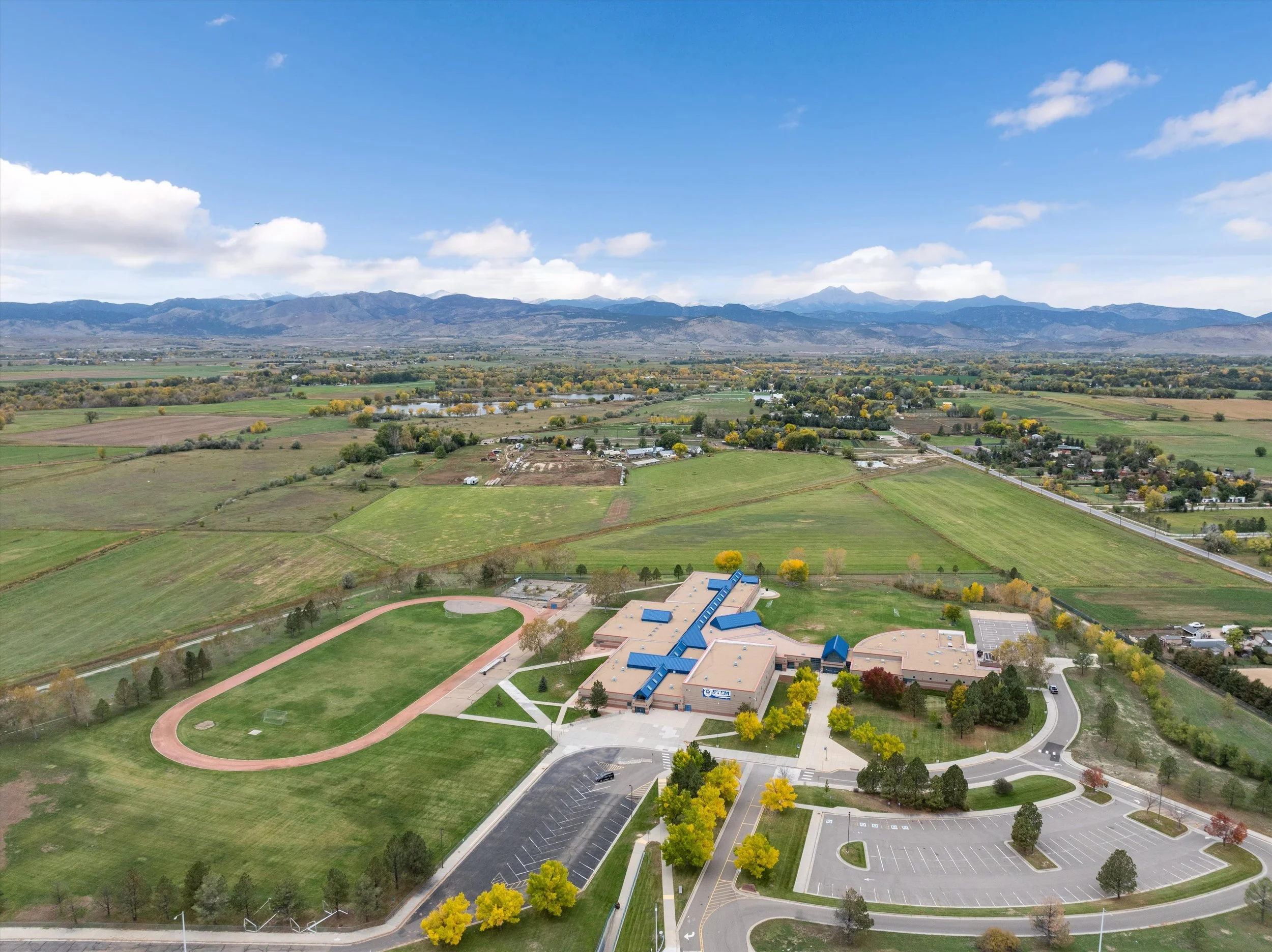 Aerial view of a Westview Middle School. Located 2 minutes from 3801 Fowler Ln Longmont Co 80503. The school is surrounded by green fields, parking lot, and sidewalks, with mountains in the background on a partly cloudy day.