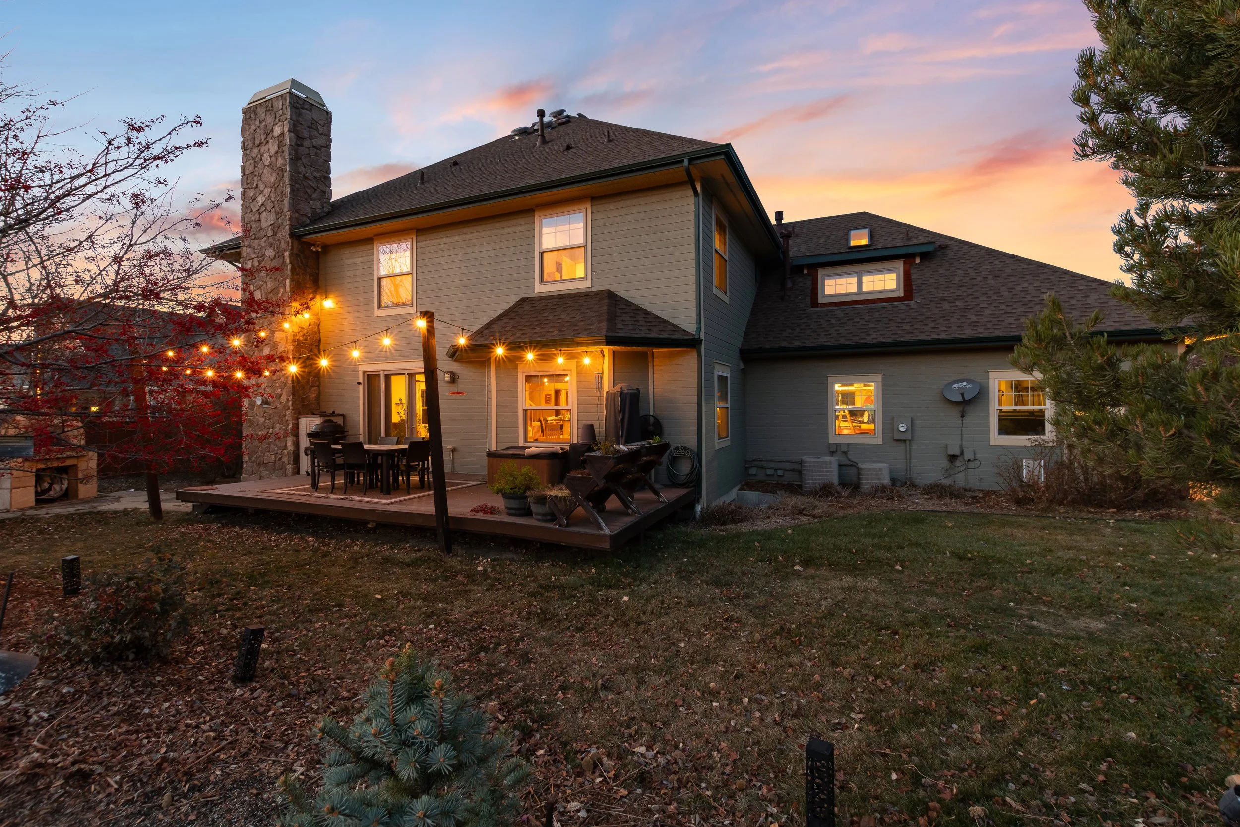 A backyard patio with string lights, outdoor pizza oven and fountain adjacent to a two-story house at sunset.