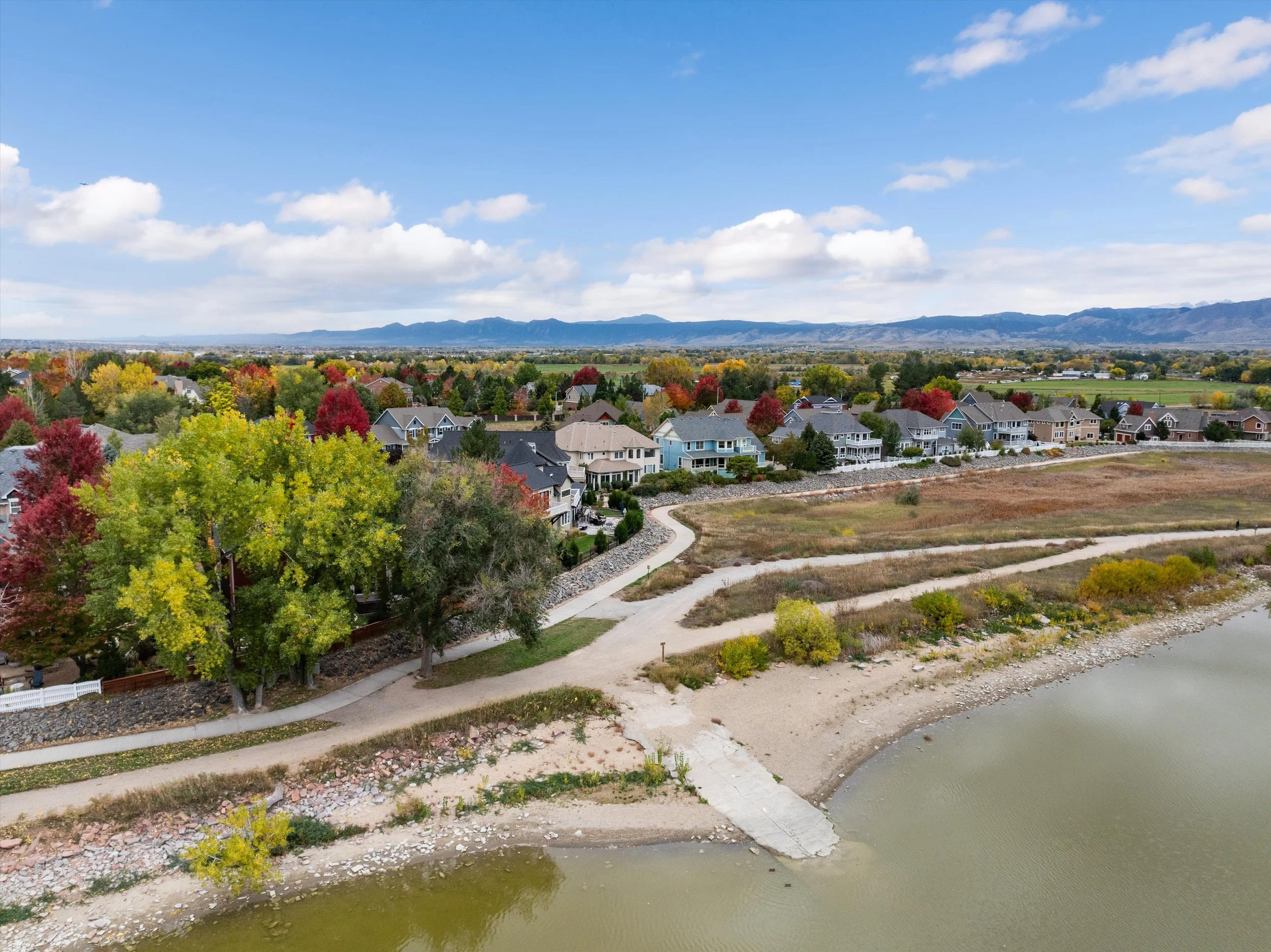 A lakeside community with a walking path, colorful trees, houses, and mountains in the background on a partly cloudy day.
