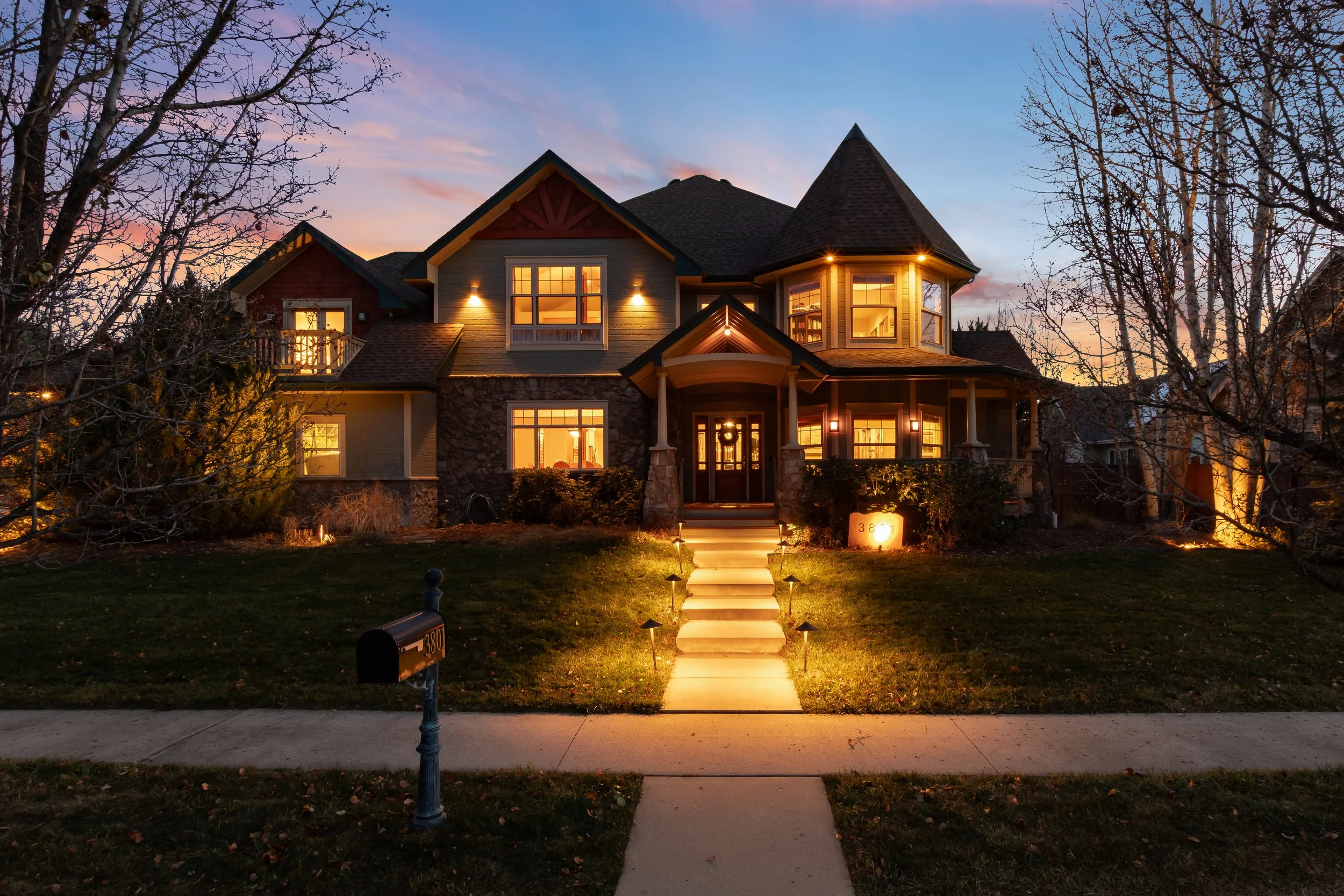 A large, two-story Victorian-style house illuminated with warm interior and exterior lights during twilight. It has a prominent turret, a front porch, and a well-maintained lawn with a lit pathway leading to the front door. The sky shows a pink and b