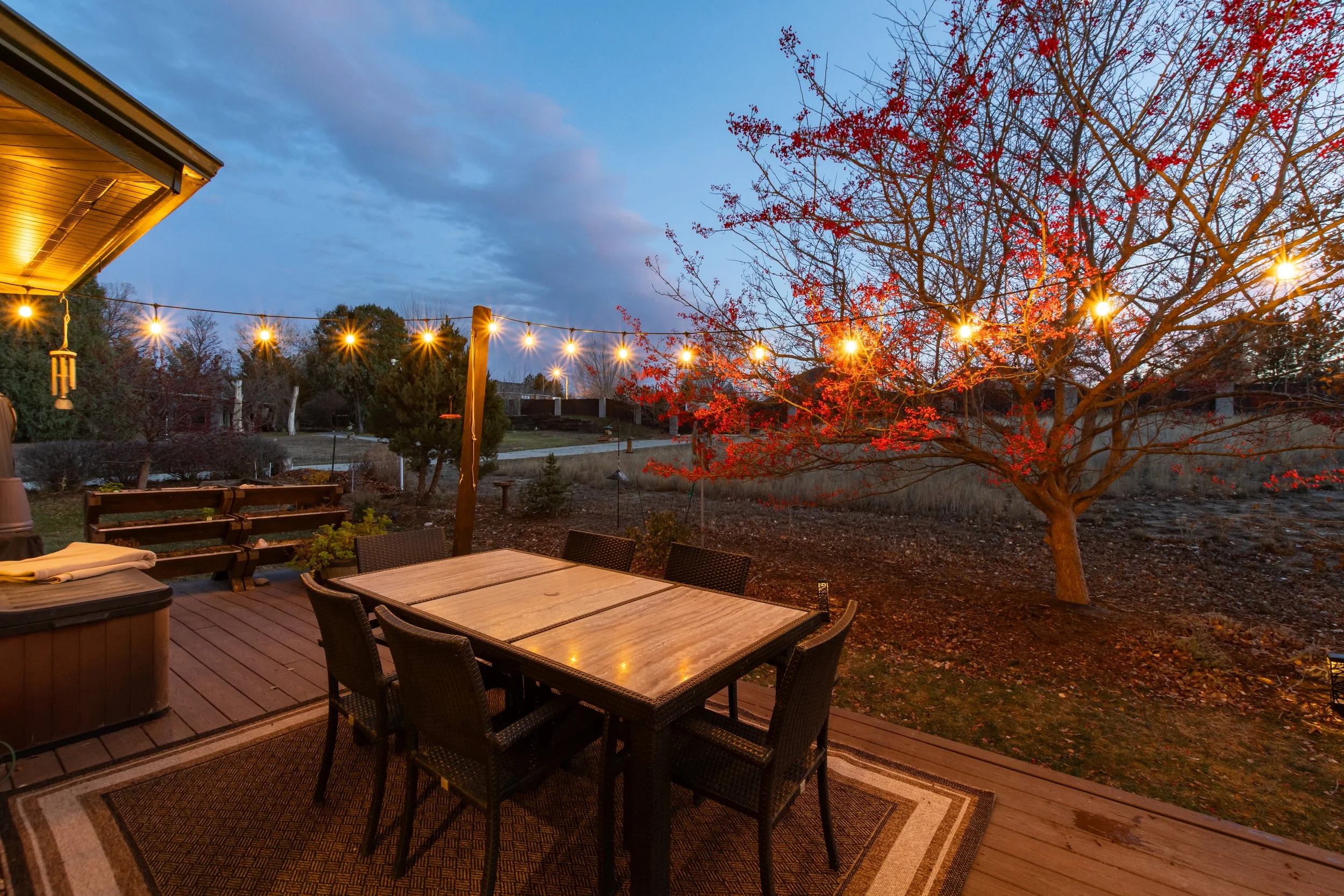 A backyard patio at dusk with a wooden table and six chairs on a rug, string lights hanging above, a tree with red leaves, and a cloudy sky in the background.