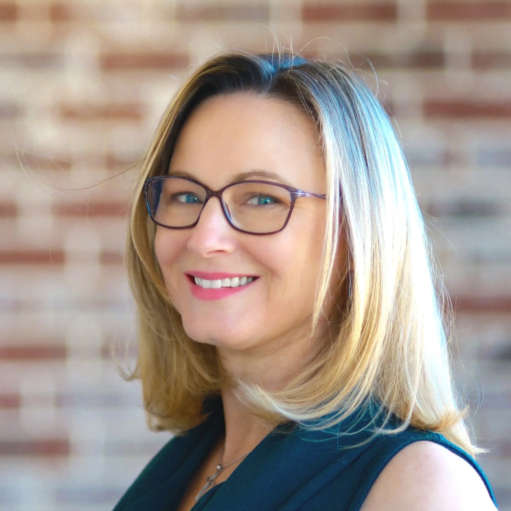 Kim Taylor, A smiling woman with blonde hair wearing glasses and a dark sleeveless top, standing in front of a brick wall.