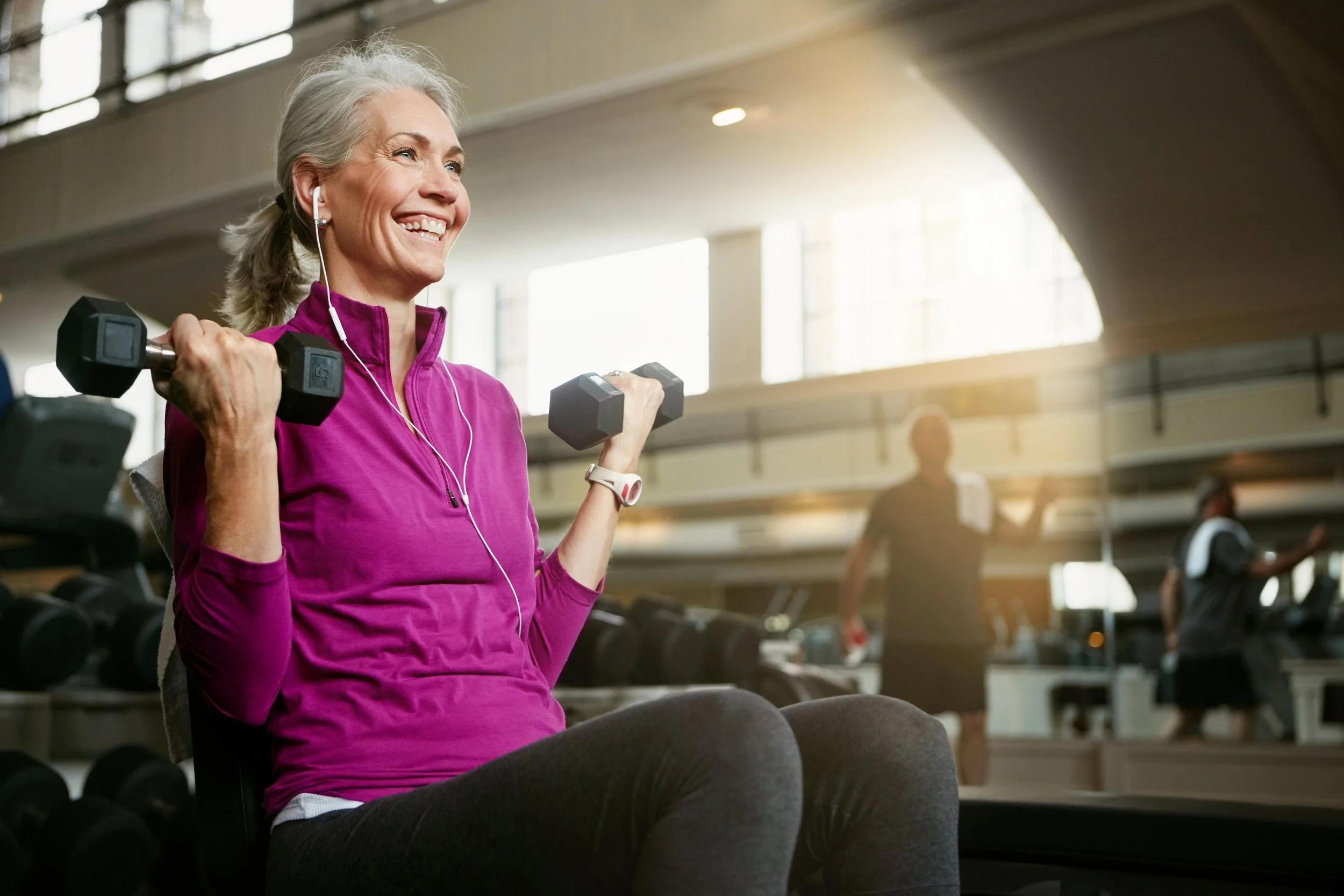 Older woman in a purple long sleeve shirt exercising with dumbbells at a gym, smiling, with headphones on, in a bright, modern fitness center.