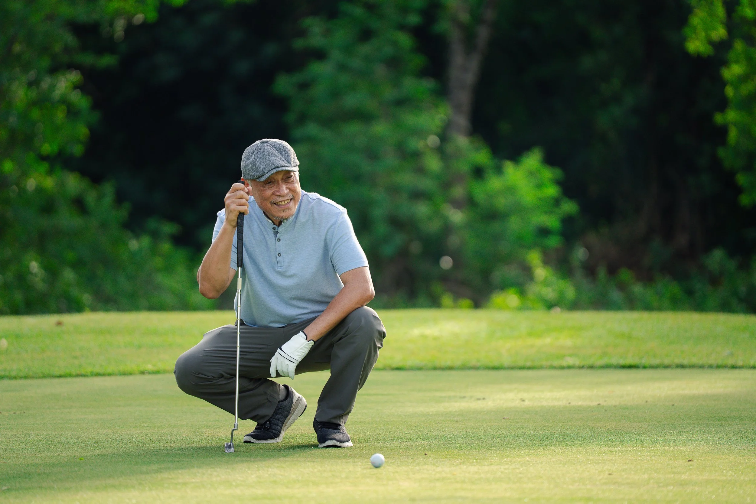Smiling elderly man with gray hair and beard, wearing a gray hat, light blue shirt, and gray pants, crouching on a golf course holding a golf club, with a golf ball in front of him and lush trees in the background.