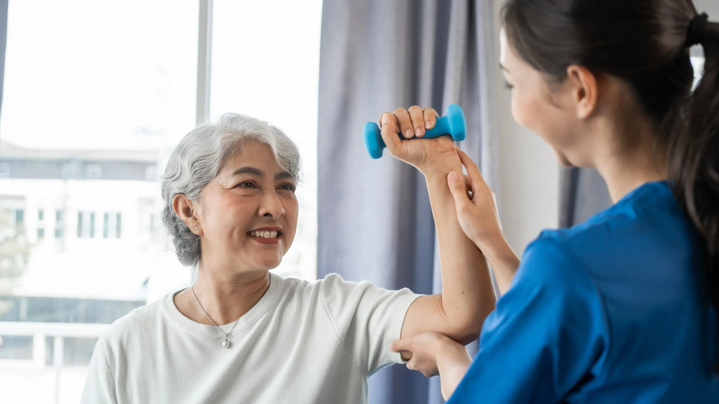 Elderly woman doing arm exercise with a blue dumbbell guided by a physical therapist in a room with large window