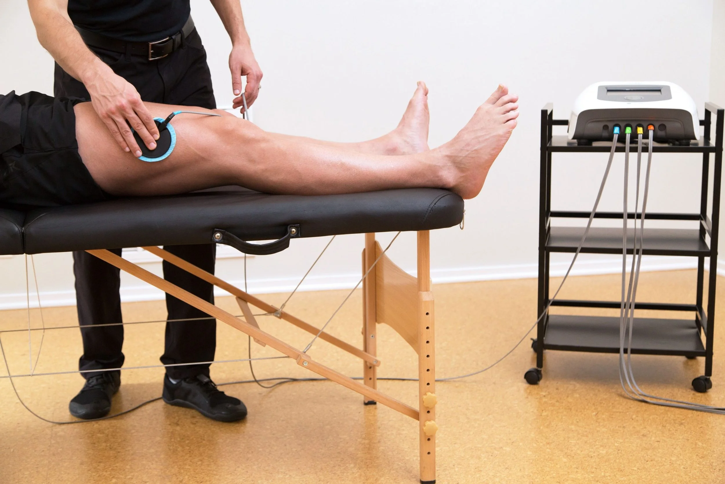 A person receiving physical therapy on their knee while lying on a black treatment table. A therapist is standing beside them, holding the person's knee, and a device connected by wires is attached to the person's knee. A medical device with multiple cables is positioned to the right on a stand.