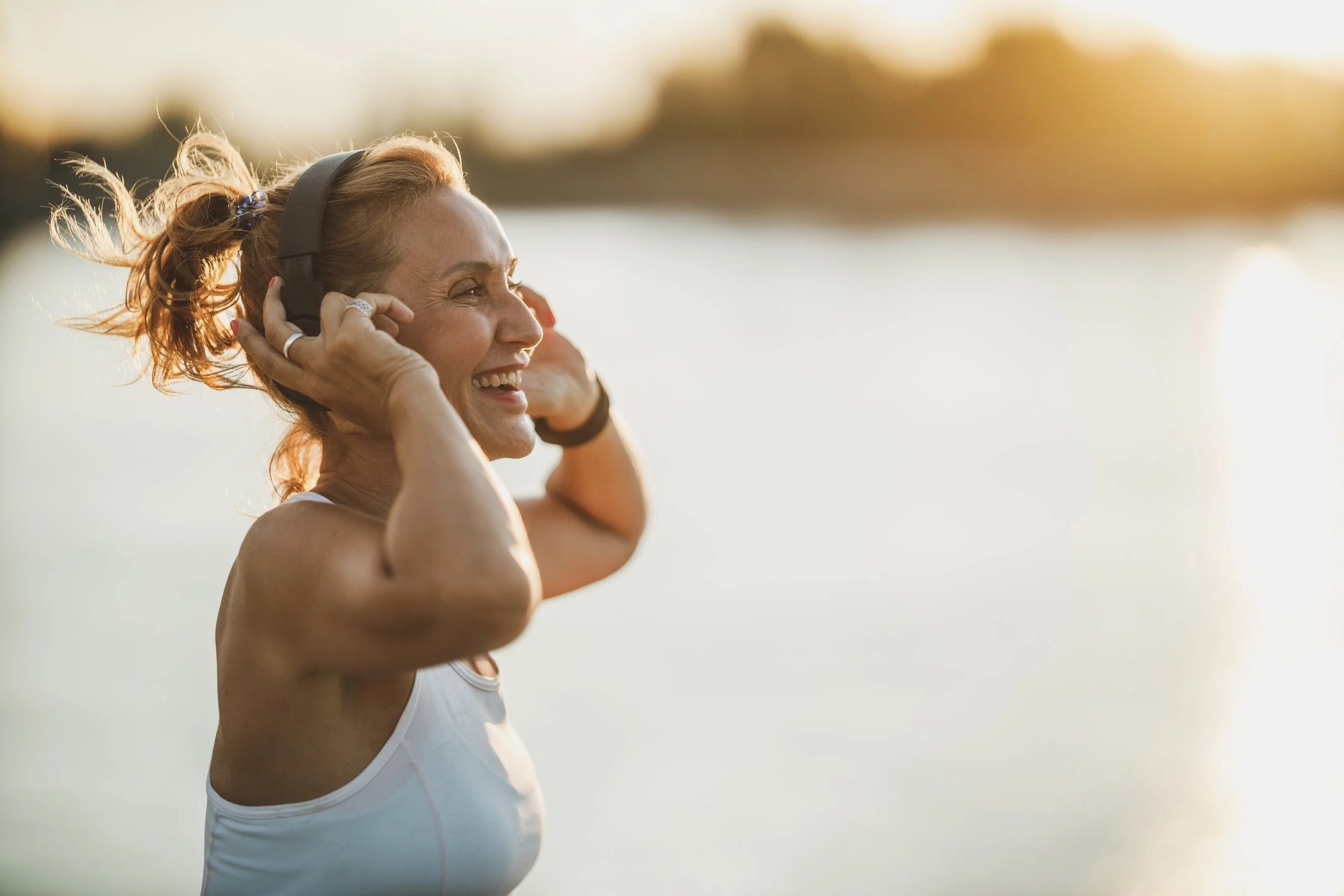 A woman with red hair tied in a bun, wearing a white sports tank top, smiling as she puts on headphones outdoors during sunset.
