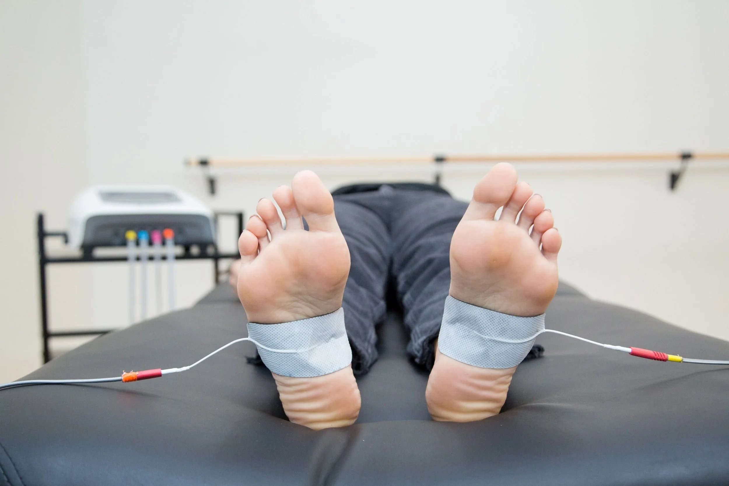 Person lying down on a medical table with electrotherapy pads attached to their feet, demonstrating a physiotherapy treatment in a clinical setting.