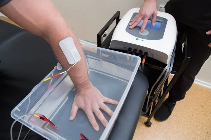 A person undergoing electrotherapy treatment with their arm submerged in a water tank connected to a medical device operated by a technician.