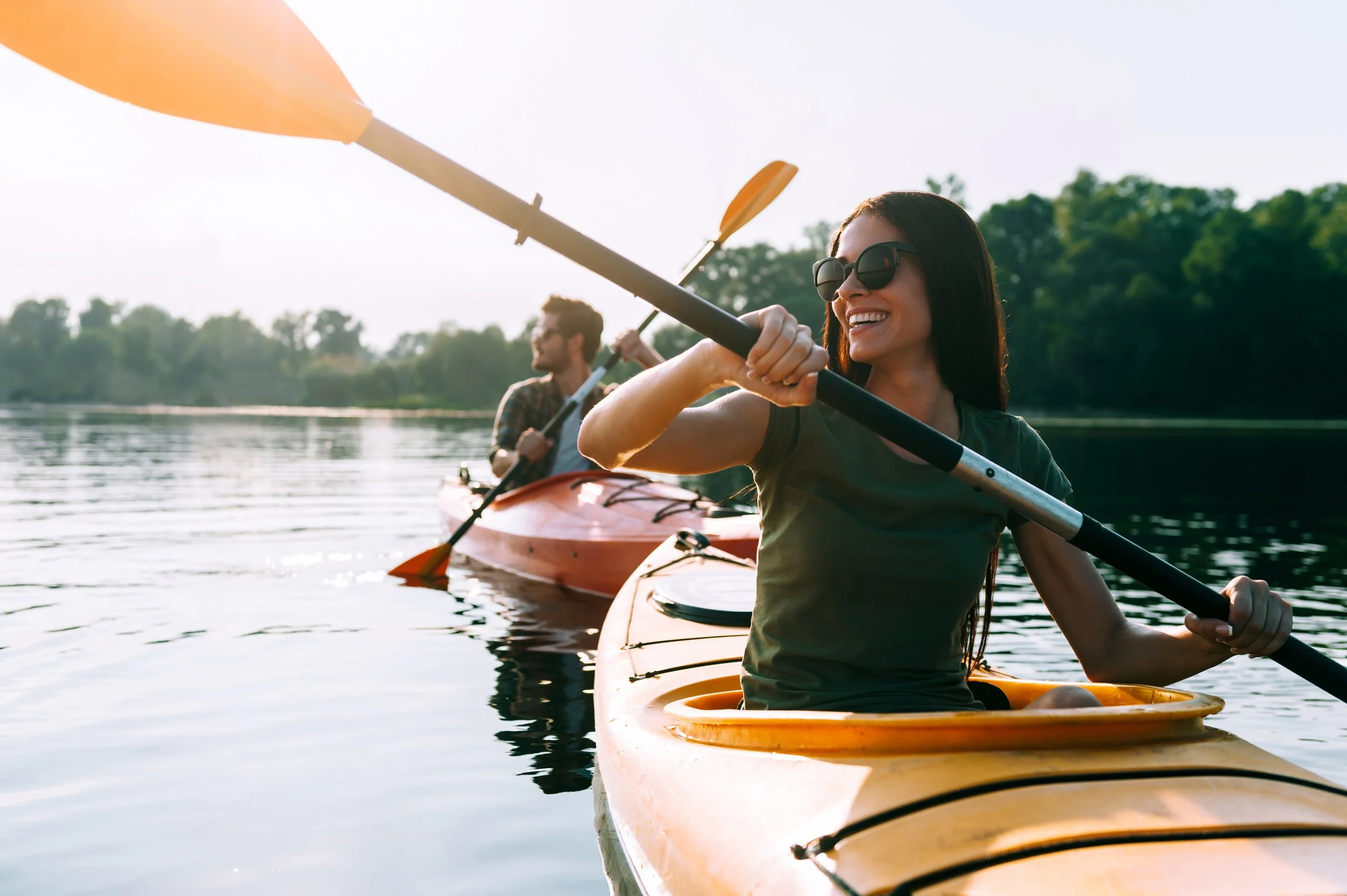 Smiling woman in sunglasses paddling a yellow kayak on a calm lake with a man in the background, surrounded by green trees.