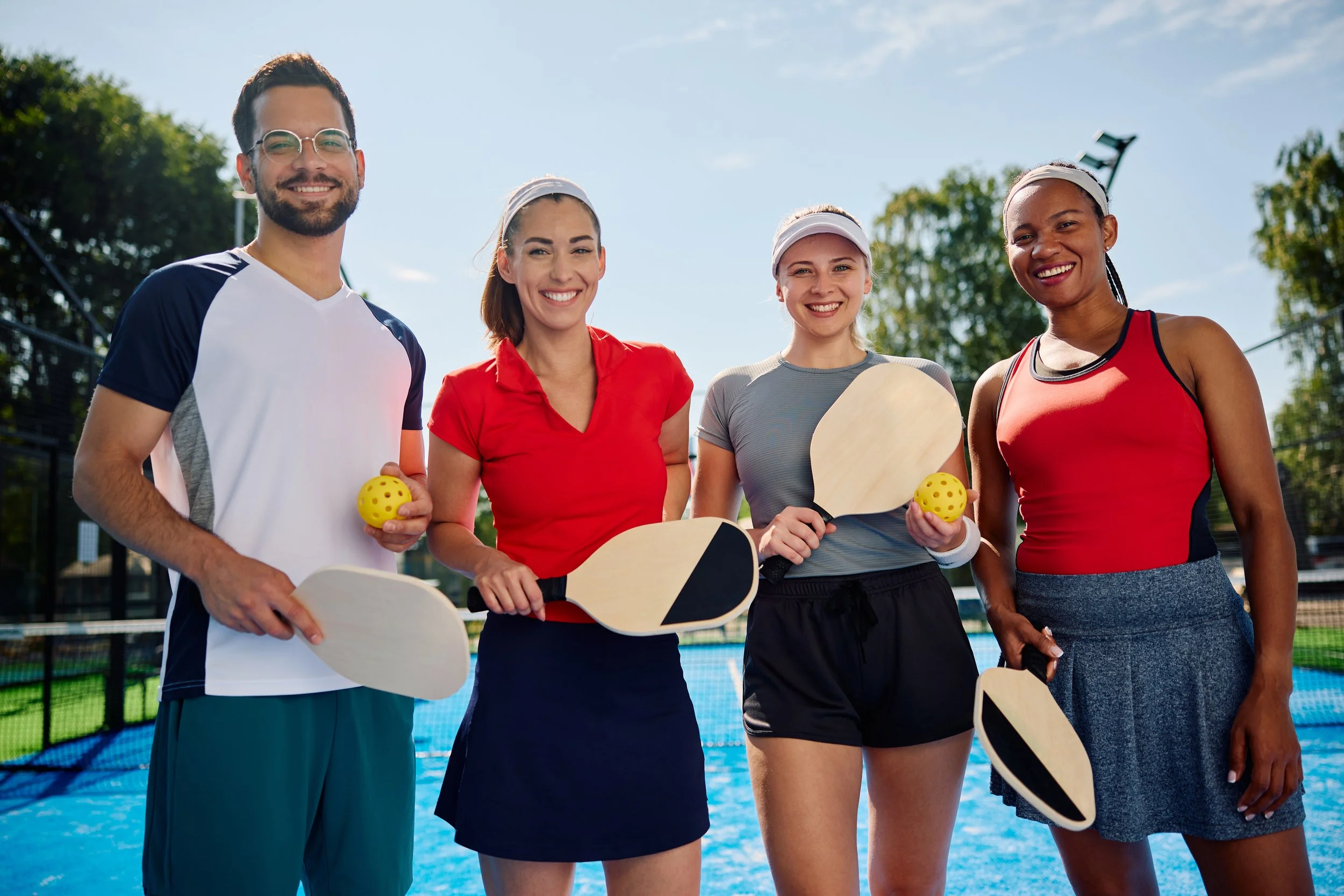 Group of four friends at a pickleball court, holding paddles and yellow pickleballs, smiling at the camera.