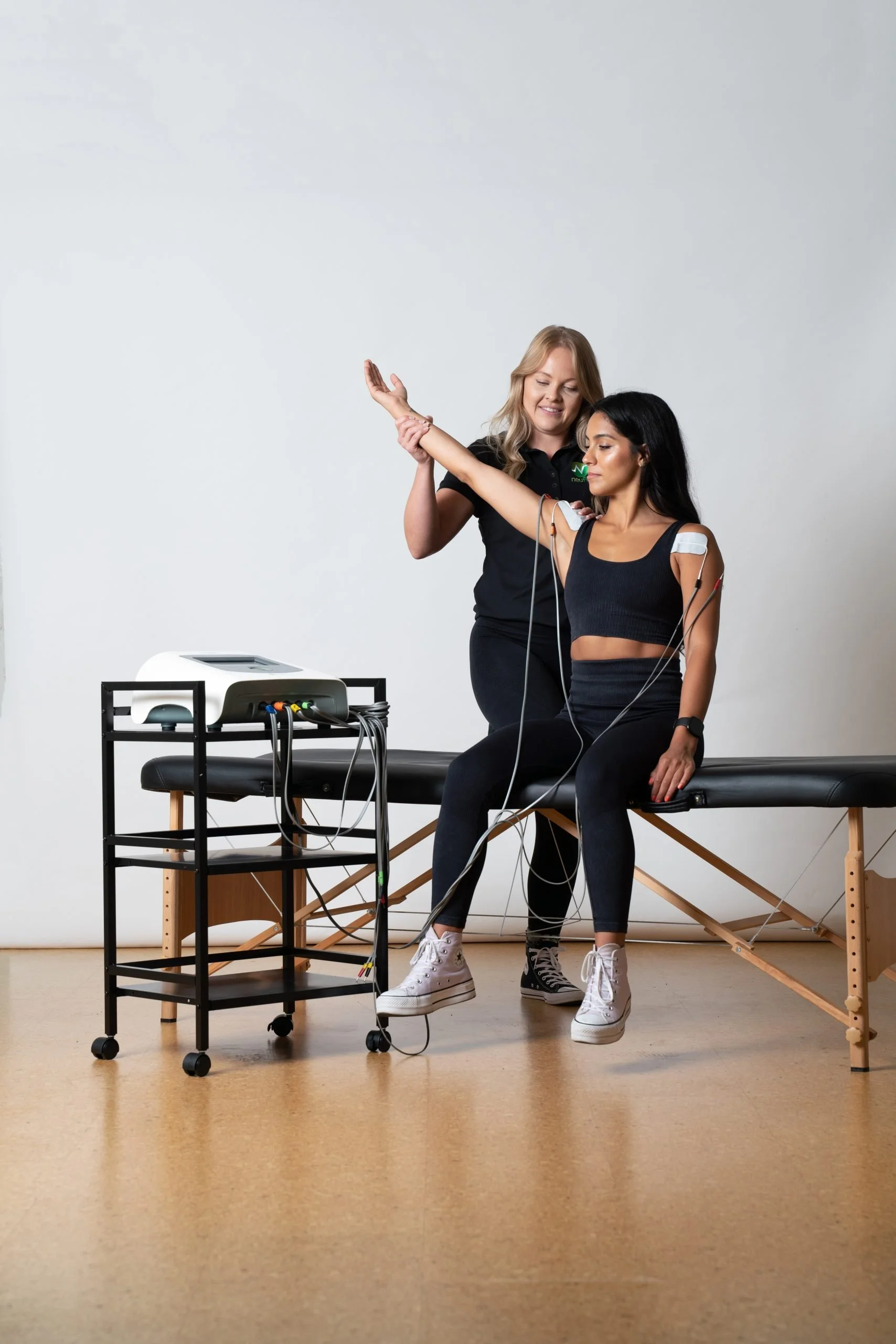 A woman with long black hair sitting on a medical examination table, having her arm tested with electrical electrodes attached. A healthcare professional stands beside her, assisting. Medical equipment and wires are nearby in a clinical setting.