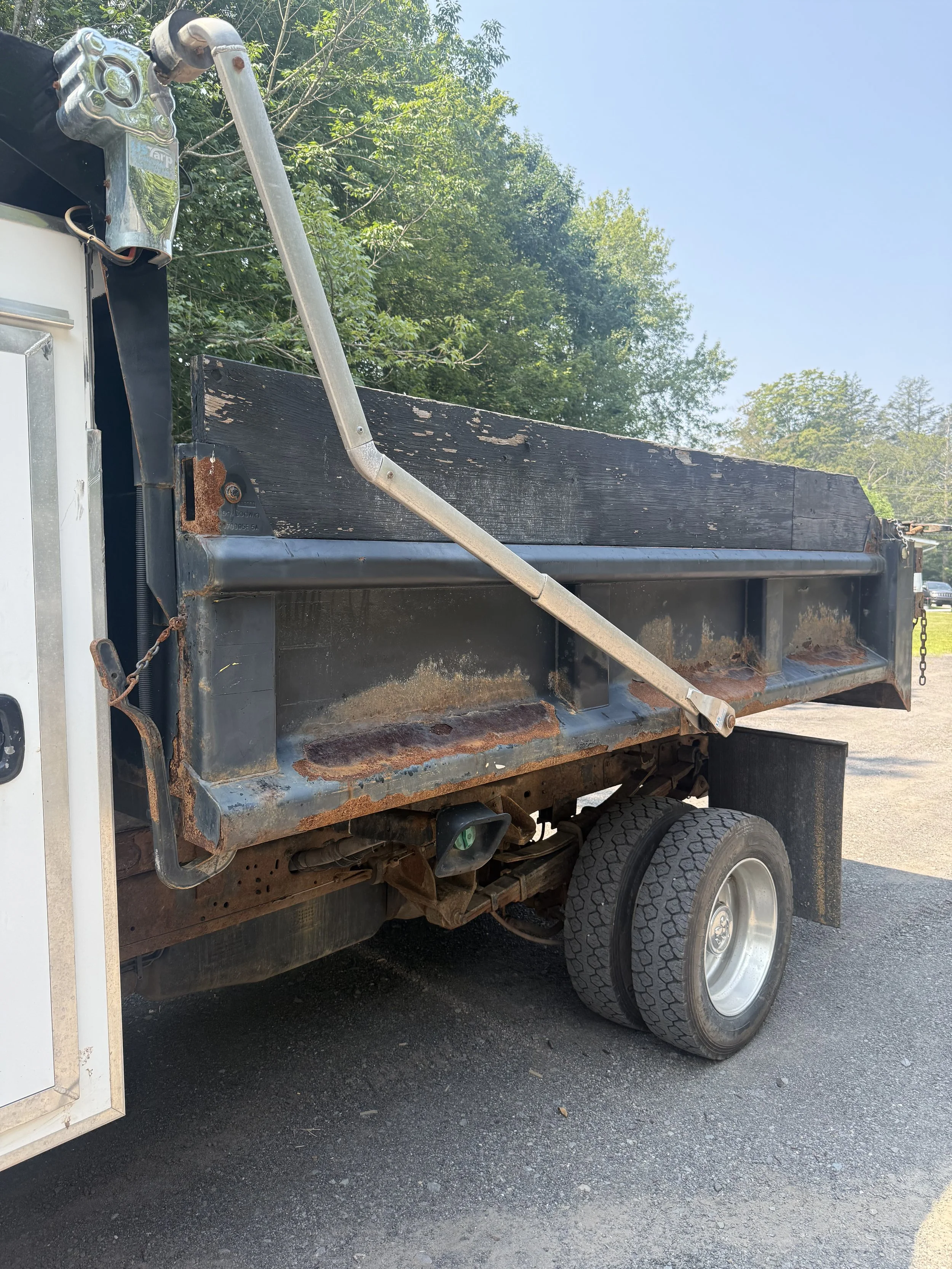 Side view of a rusty flatbed utility truck with black and silver tractor equipment, mounted on a black asphalt road with trees and blue sky in the background.