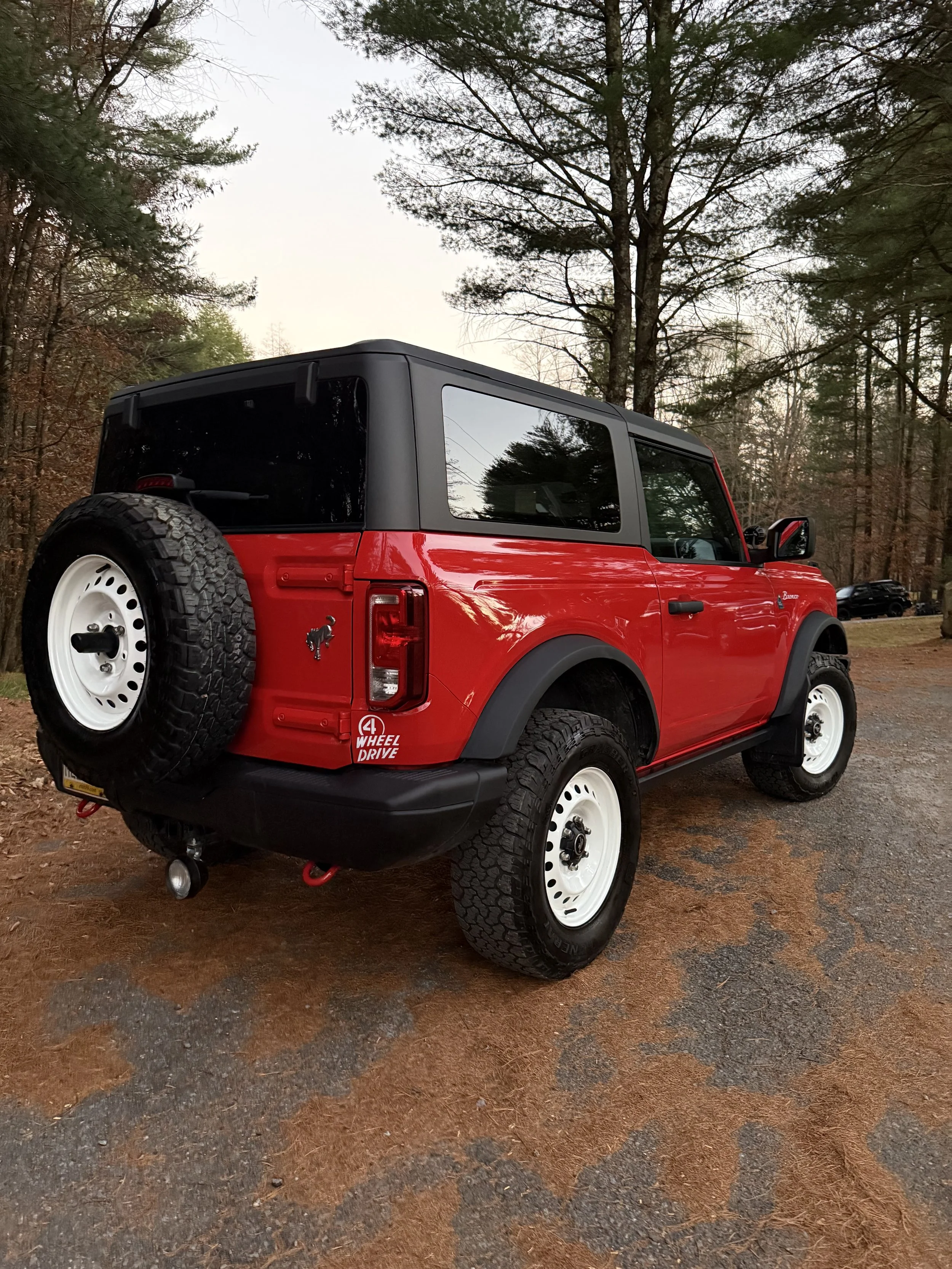 A red and black Ford Bronco with white wheels parked on a dirt path in a wooded area.