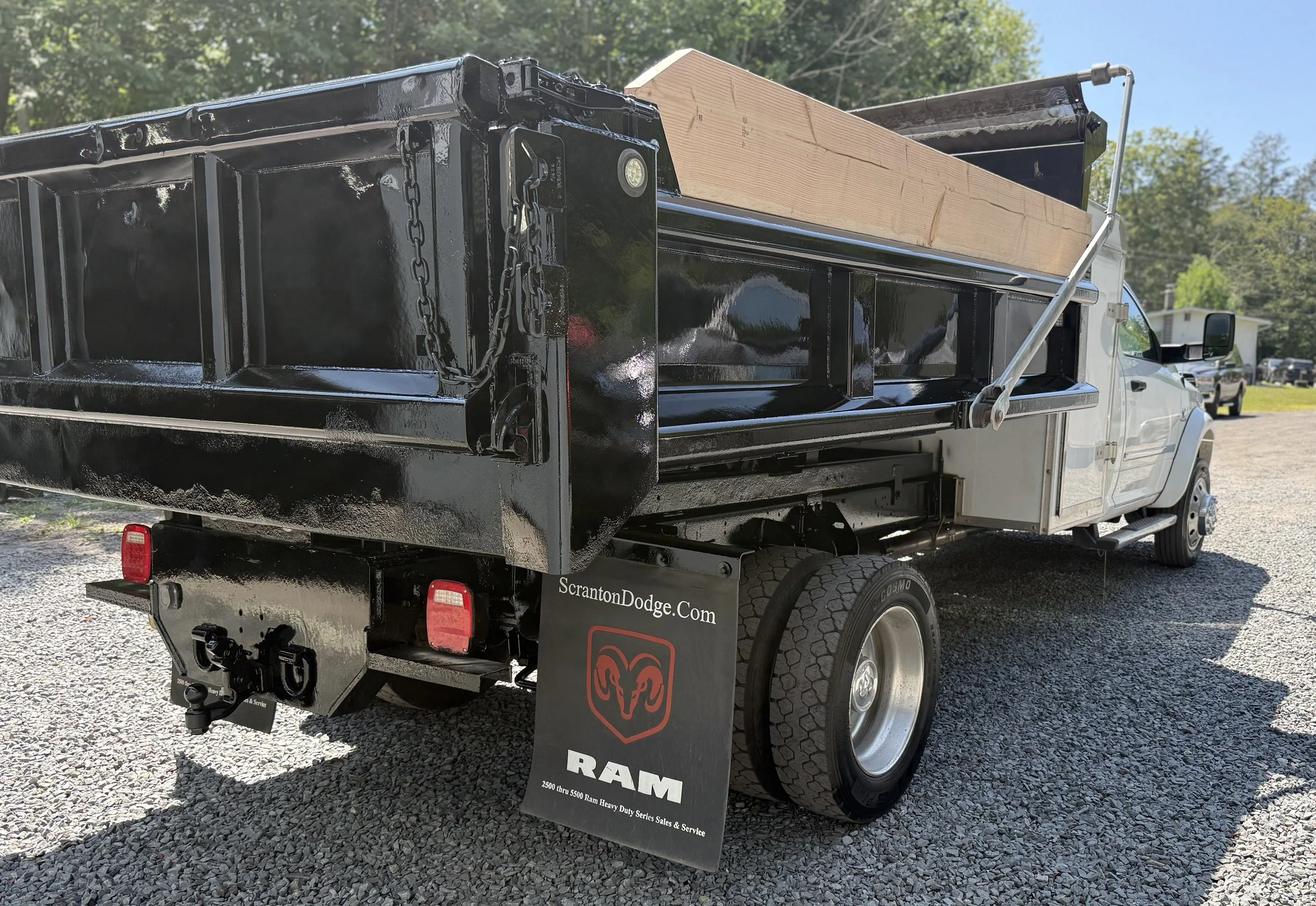 A flatbed truck with a black bed carrying a large piece of wood. The truck has a RAM sign and a website, ScrantonDodge.com, displayed on a signboard hanging from the side. The truck is parked on a gravel surface with a building and some trees visible