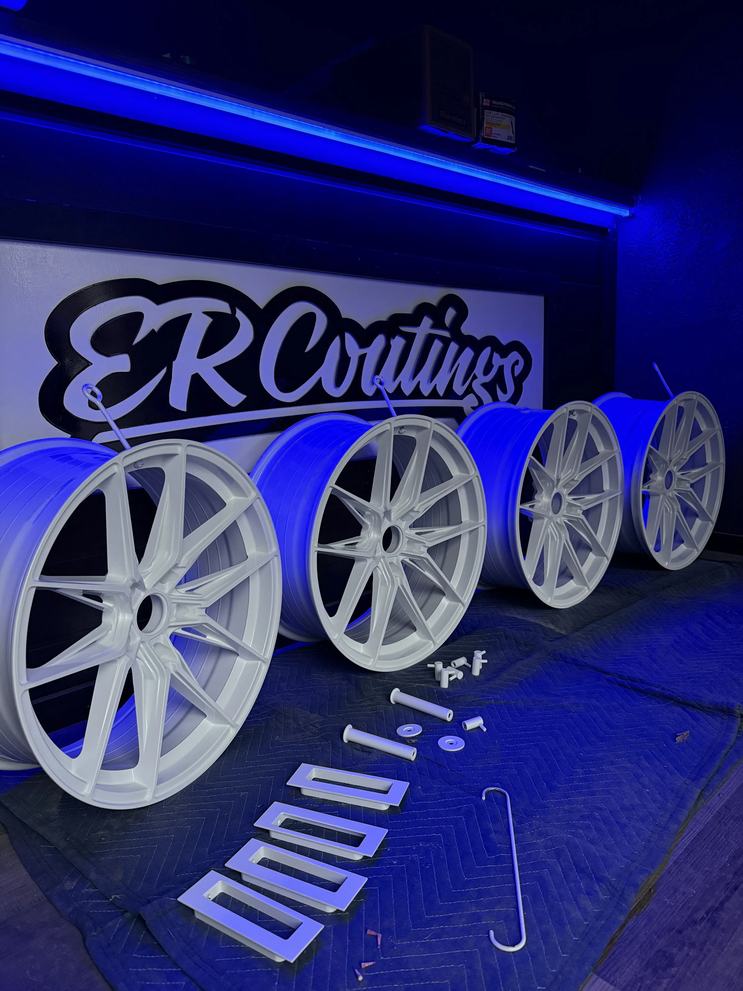 Four white car wheels with blue accents, assembly parts, and tools on a black padded surface with a sign that reads ER Couture in the background.