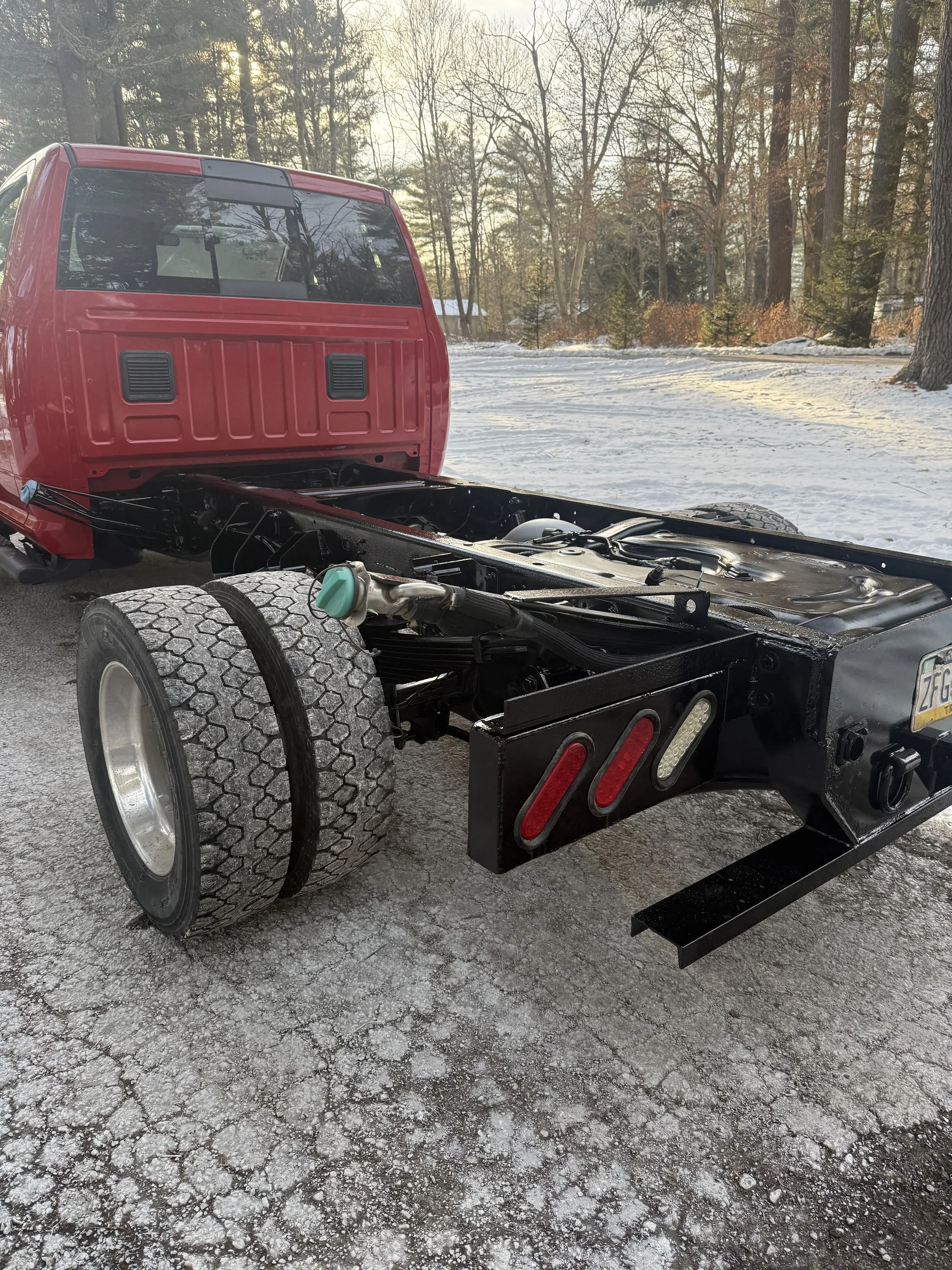 A partially assembled red truck with no cargo bed, parked on a snowy driveway surrounded by trees, in a winter landscape.