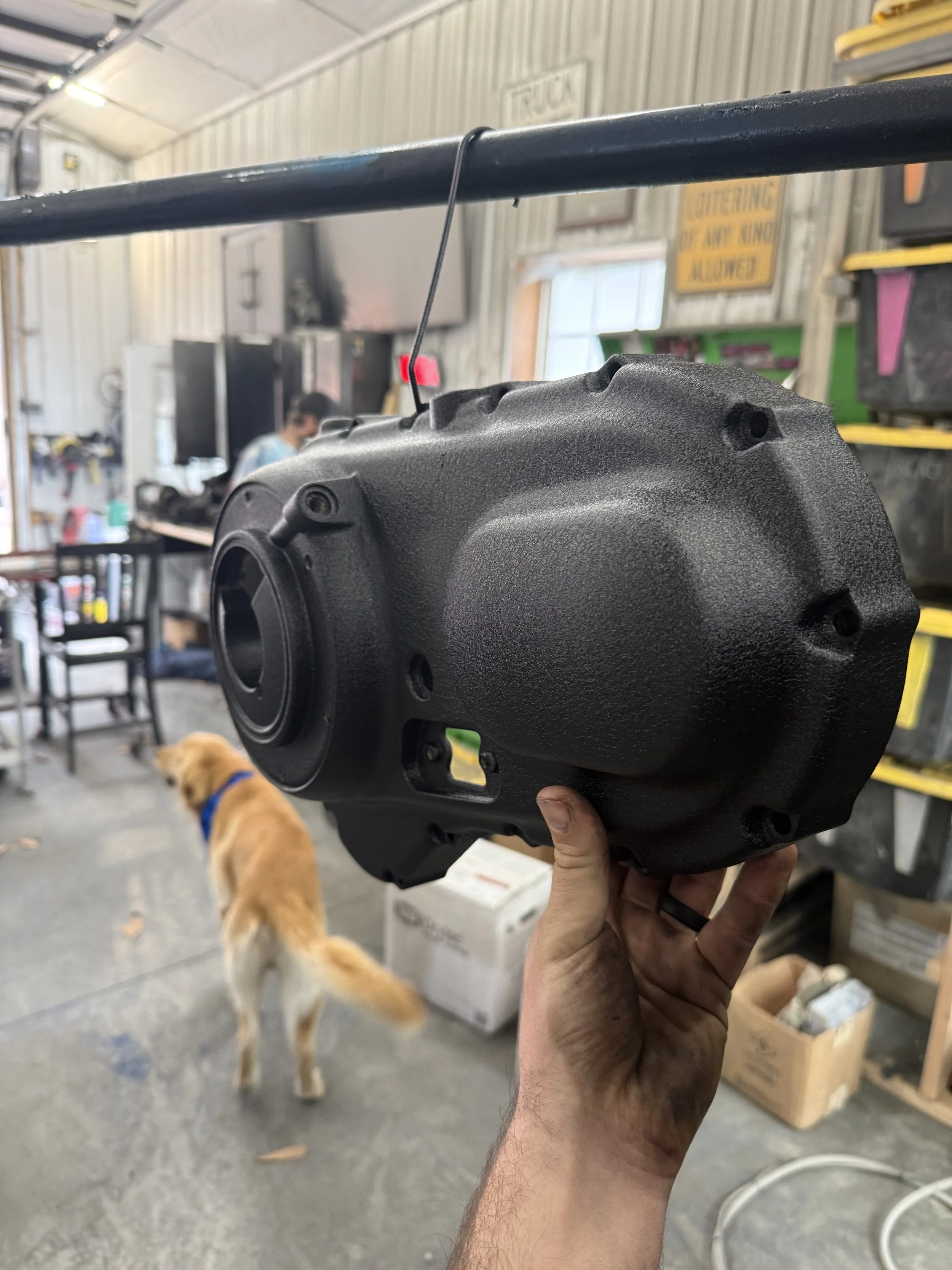Person holding a black, textured metal automotive part inside a workshop with tools, a dog, and workers in the background.