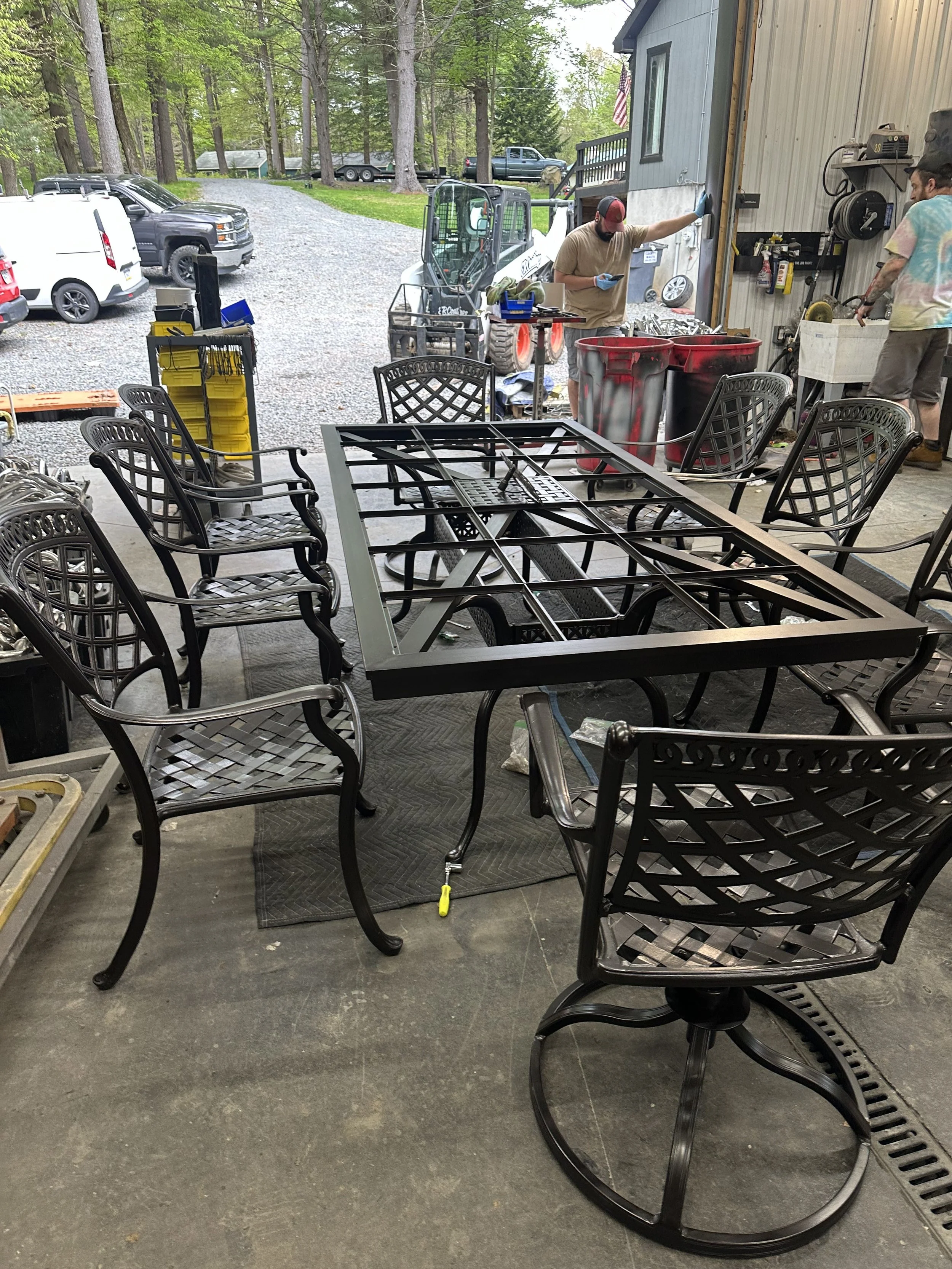 Workshop with black metal patio furniture and a metal frame on the table in the center, two men working outside near a small construction vehicle, with trees and parked cars in the background.