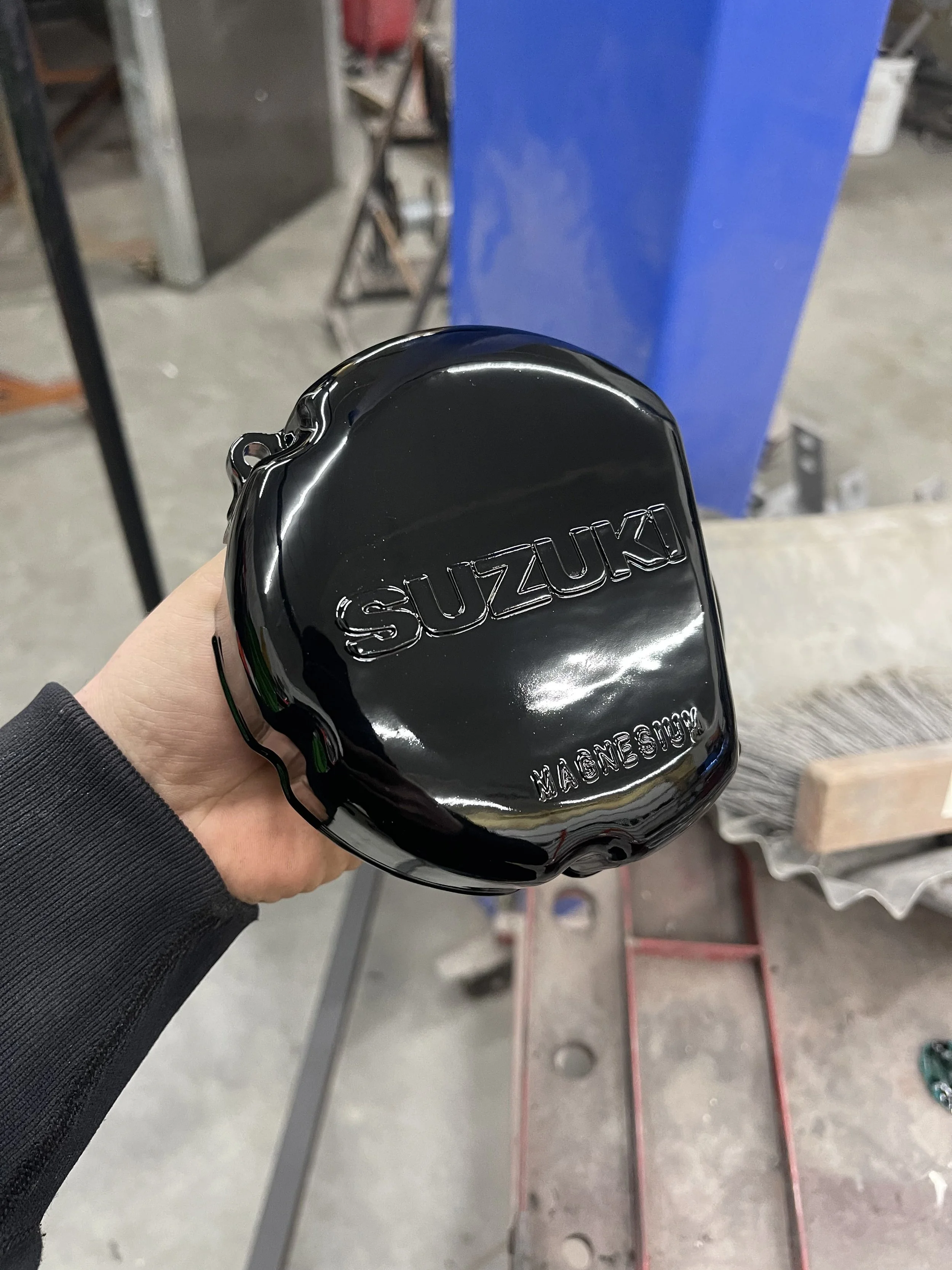 A shiny black Suzuki engine cover held by a person's hand, with the word 'SUZUKI' embossed on it and 'MAGNEFLOW' written below.
